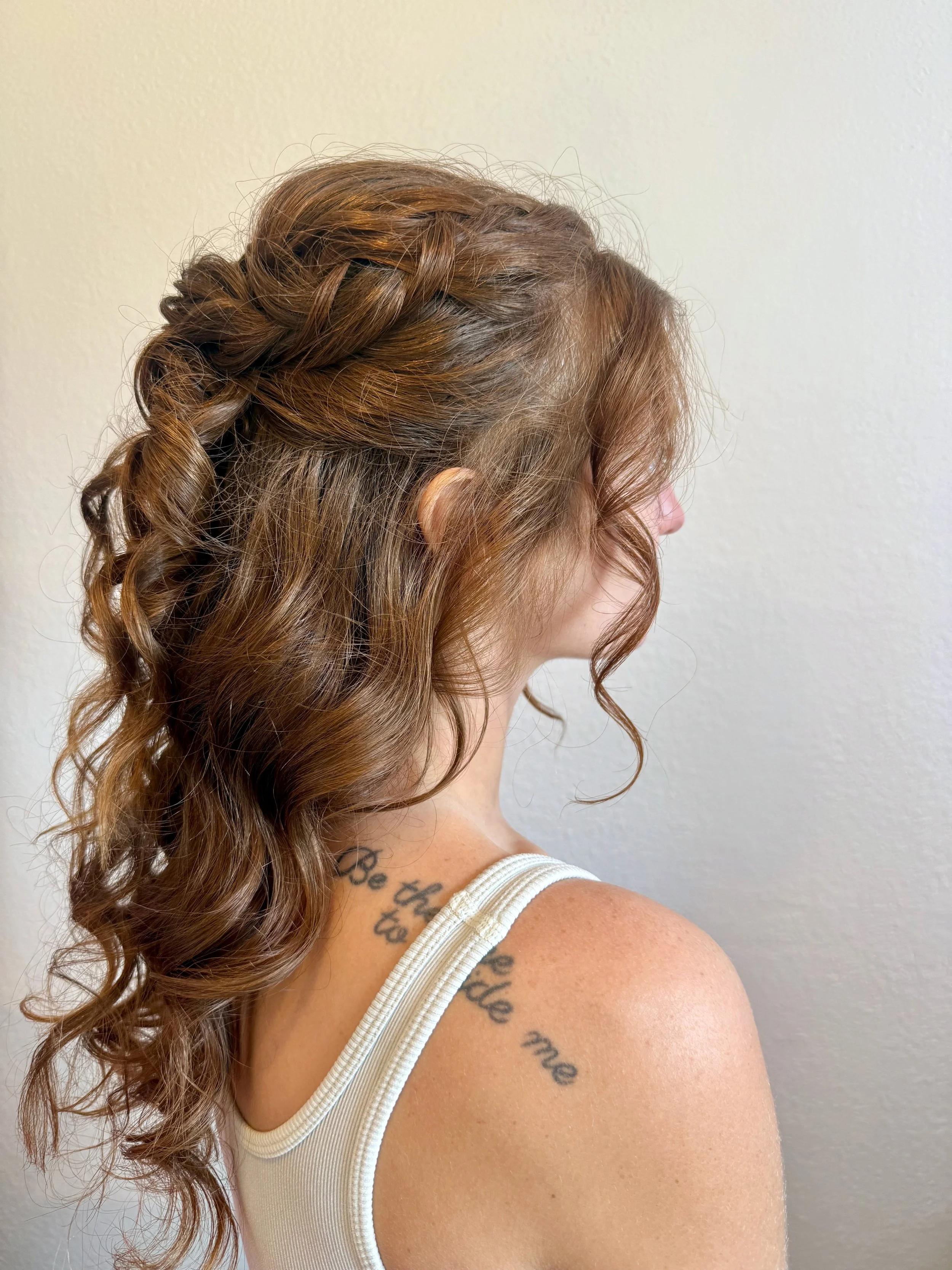 Side view of a bride with curly brown hair styled in a braid and loose curls, wearing a white tank top, showing a tattoo on her shoulder and upper back, standing against a plain wall.