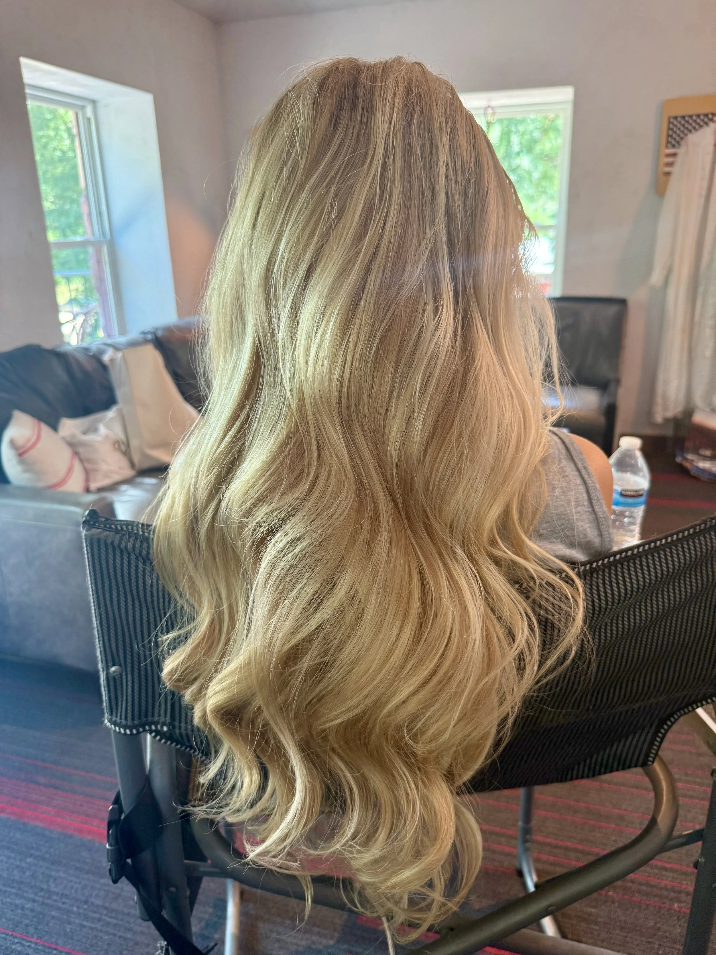 Blonde hair with loose waves draping over a black mesh office chair in a room with windows, a couch, and a water bottle on a table.