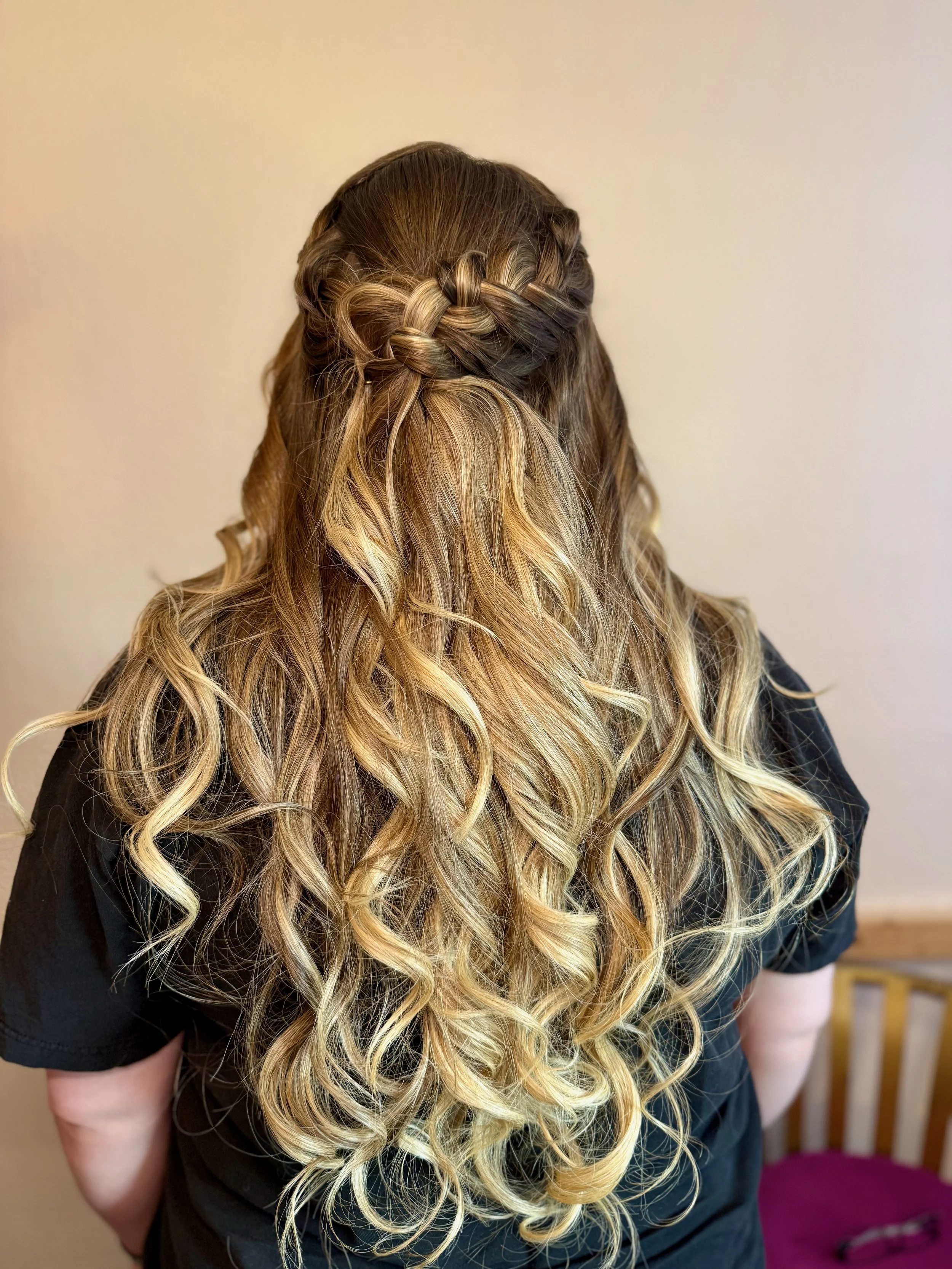 Back view of a bride with long, wavy hair featuring a braided crown at the top, blending dark brown and blonde hair.