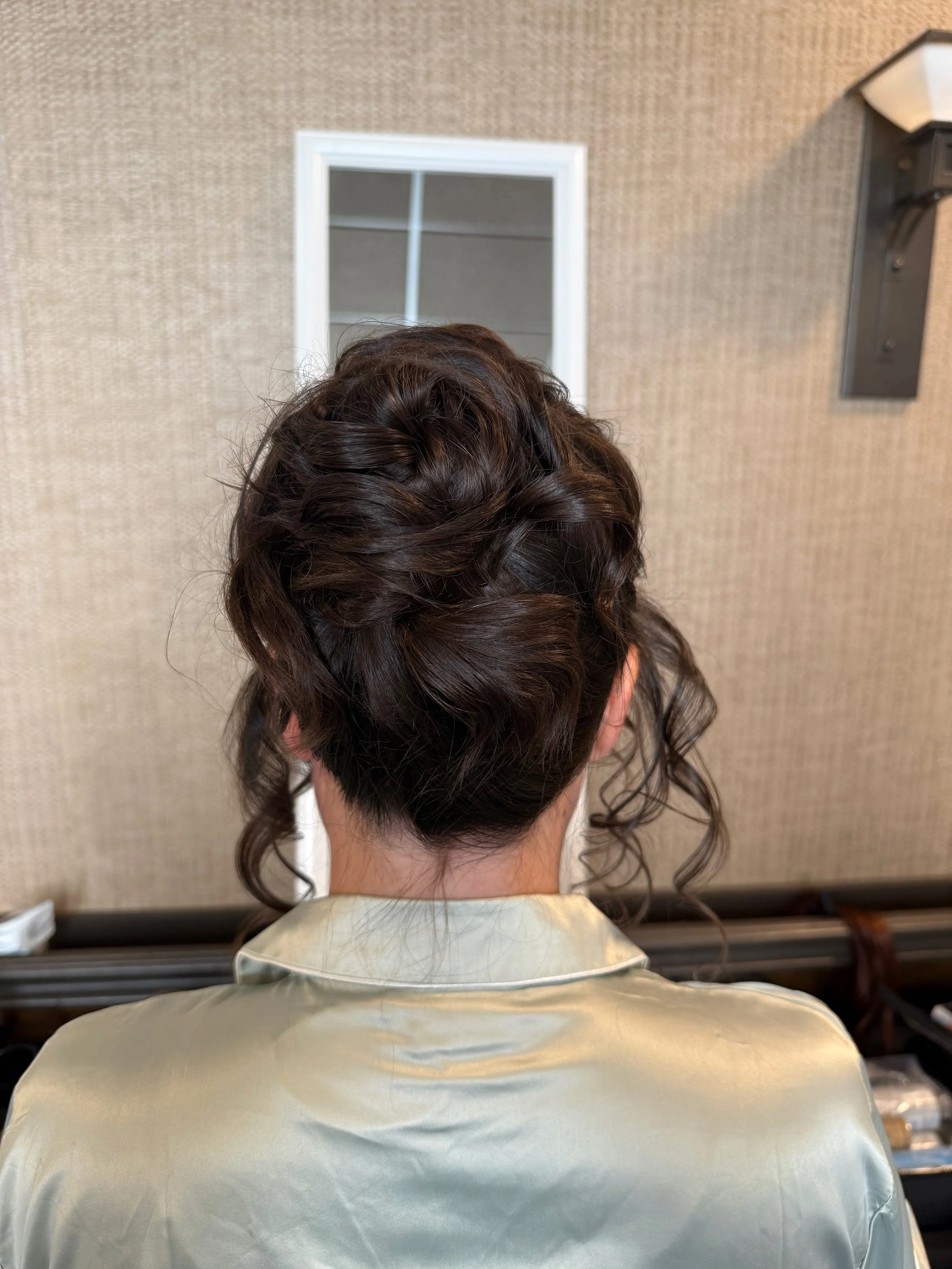 Back of a bride with styled, curly brown hair, sitting in a salon chair, wearing a beige cape, with a mirror on the wall behind her.