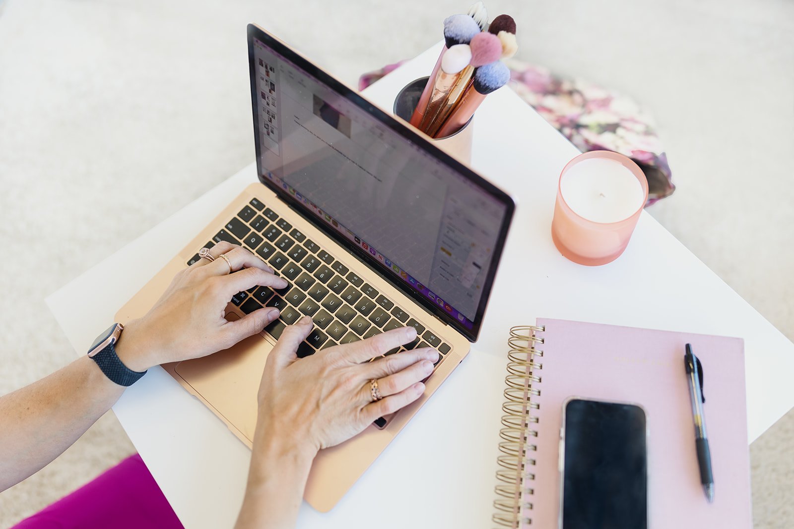 A person typing on a rose gold laptop at a white desk, with a pink notebook, a black pen, a smartphone, a pink candle, and a pink pen holder with makeup brushes.