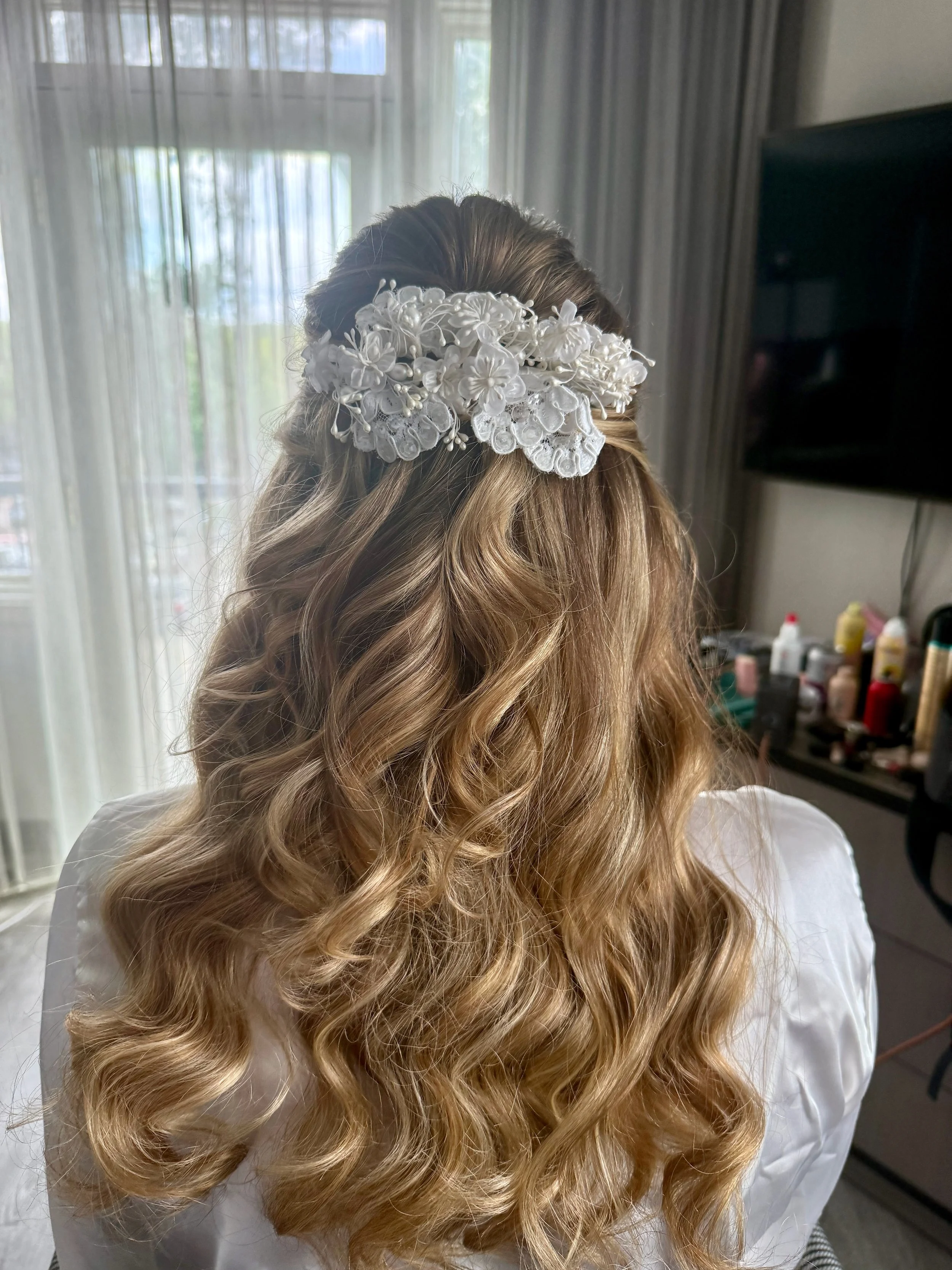 A bride with long, wavy, blonde hair wears a white floral hair accessory with fabric and pearl details.