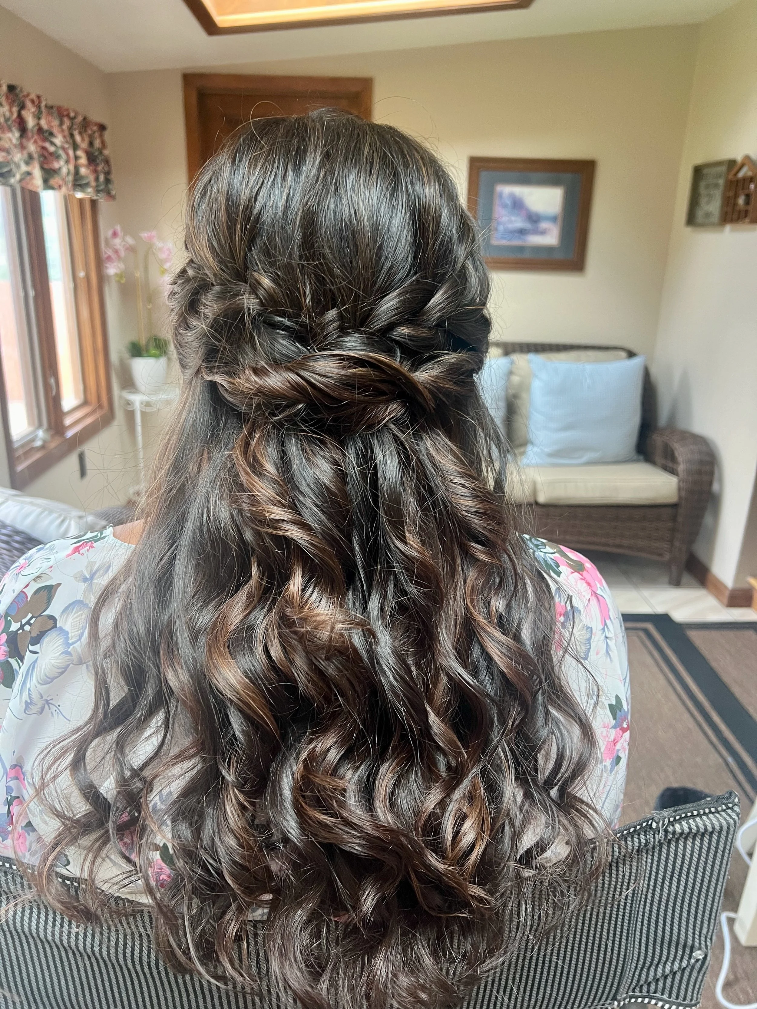 Back view of a bride with long, wavy hair styled in a half-up braid and loose curls, sitting in a room with a sofa and framed pictures on the wall.