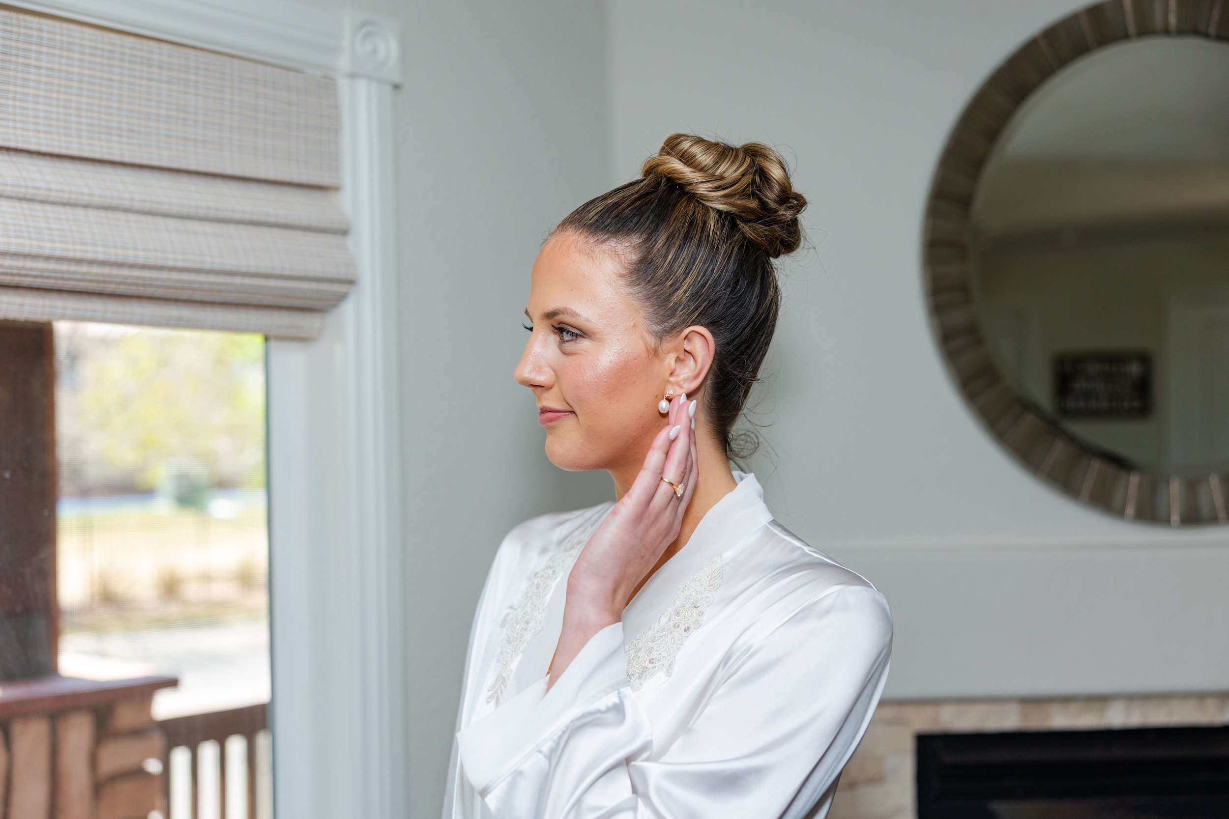 A woman with up-styled hair, pearl earrings, and a white satin robe is adjusting her earring while looking into a mirror.