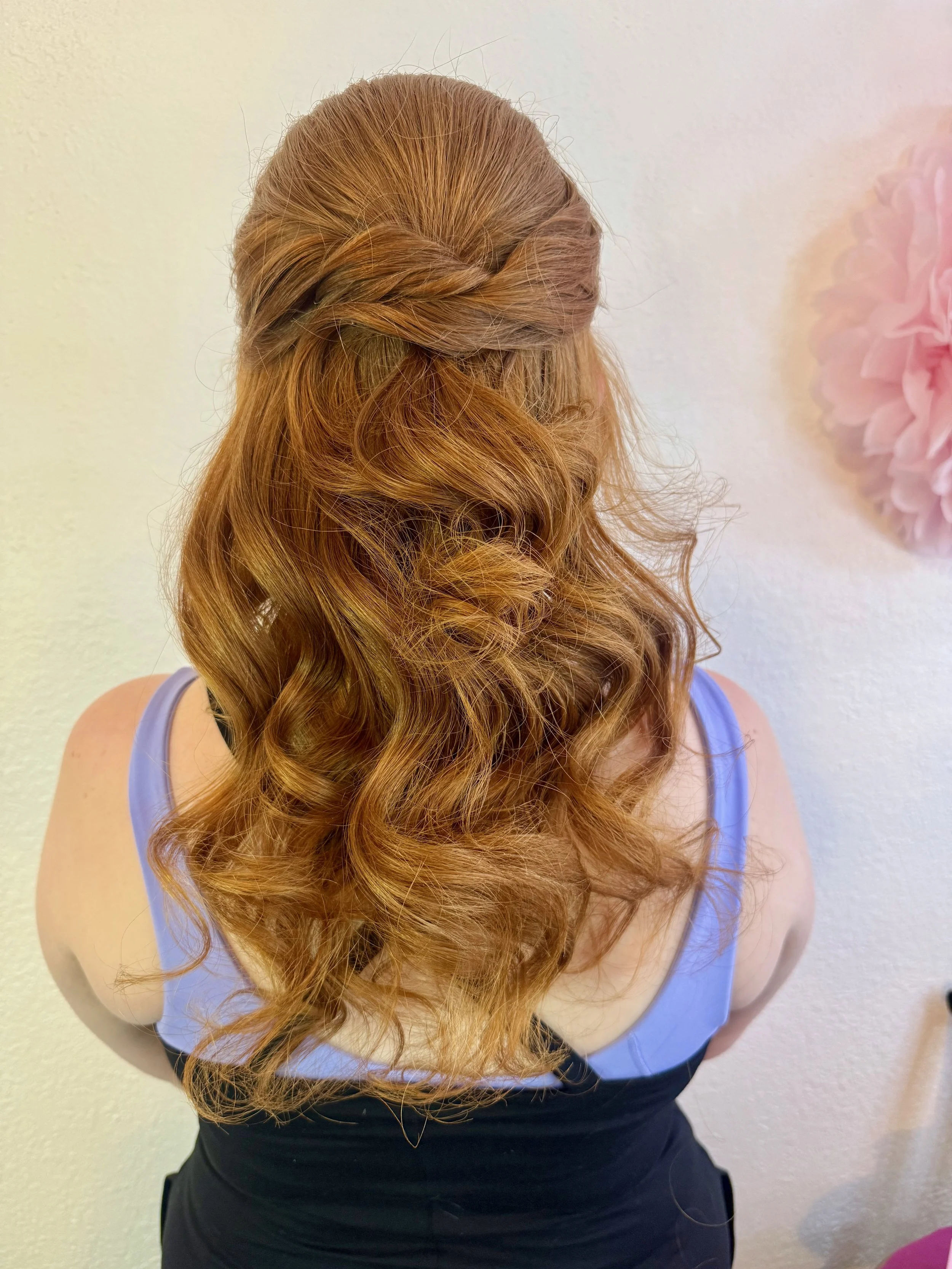 A bride with long, wavy red hair styled in loose curls, wearing a light blue sleeveless top, viewed from behind, standing in front of a plain wall with a pink flower decoration on the right side.