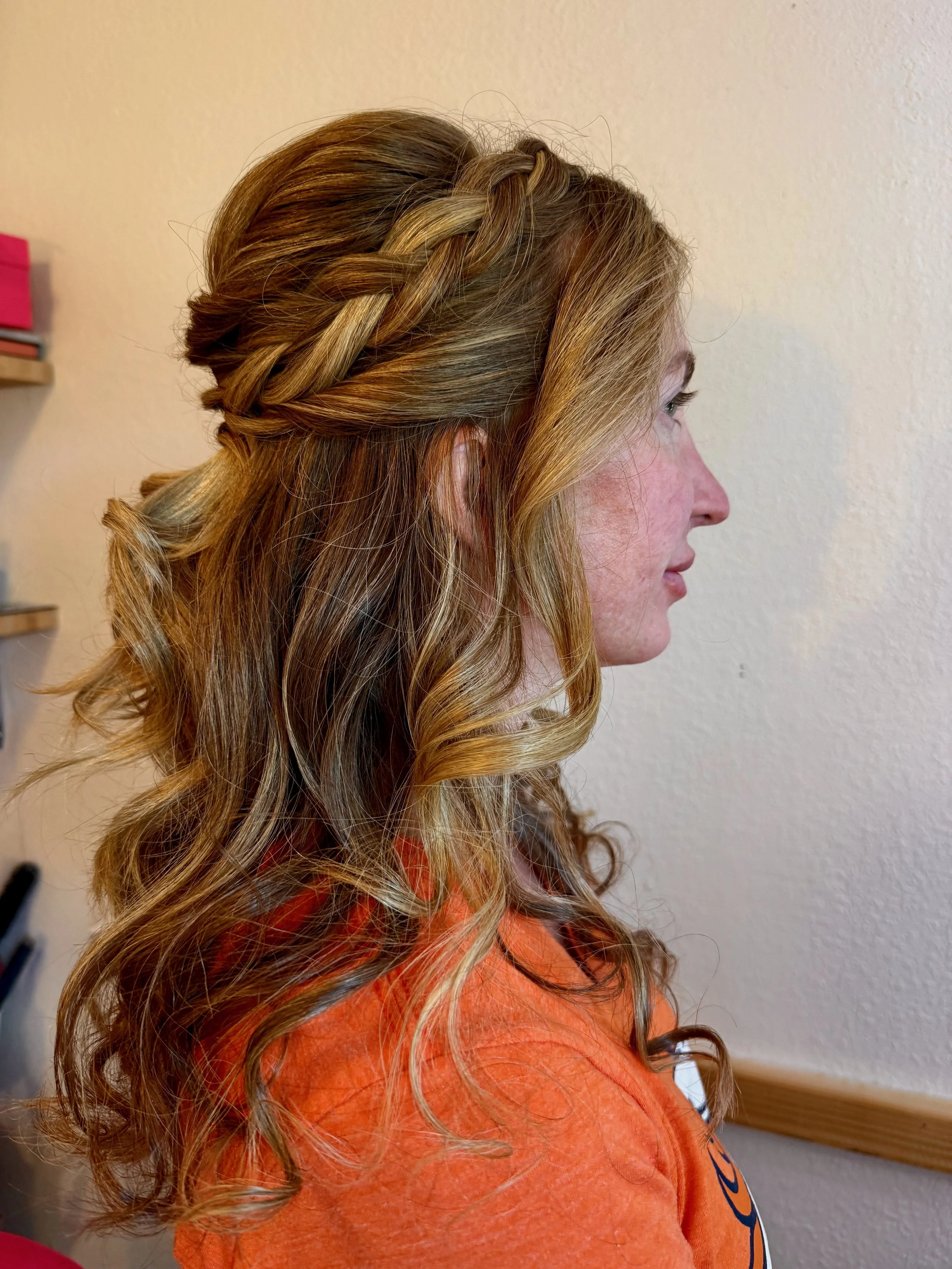 A bride with wavy blonde hair styled in a side braid, wearing an orange shirt, facing to the right against a plain wall.