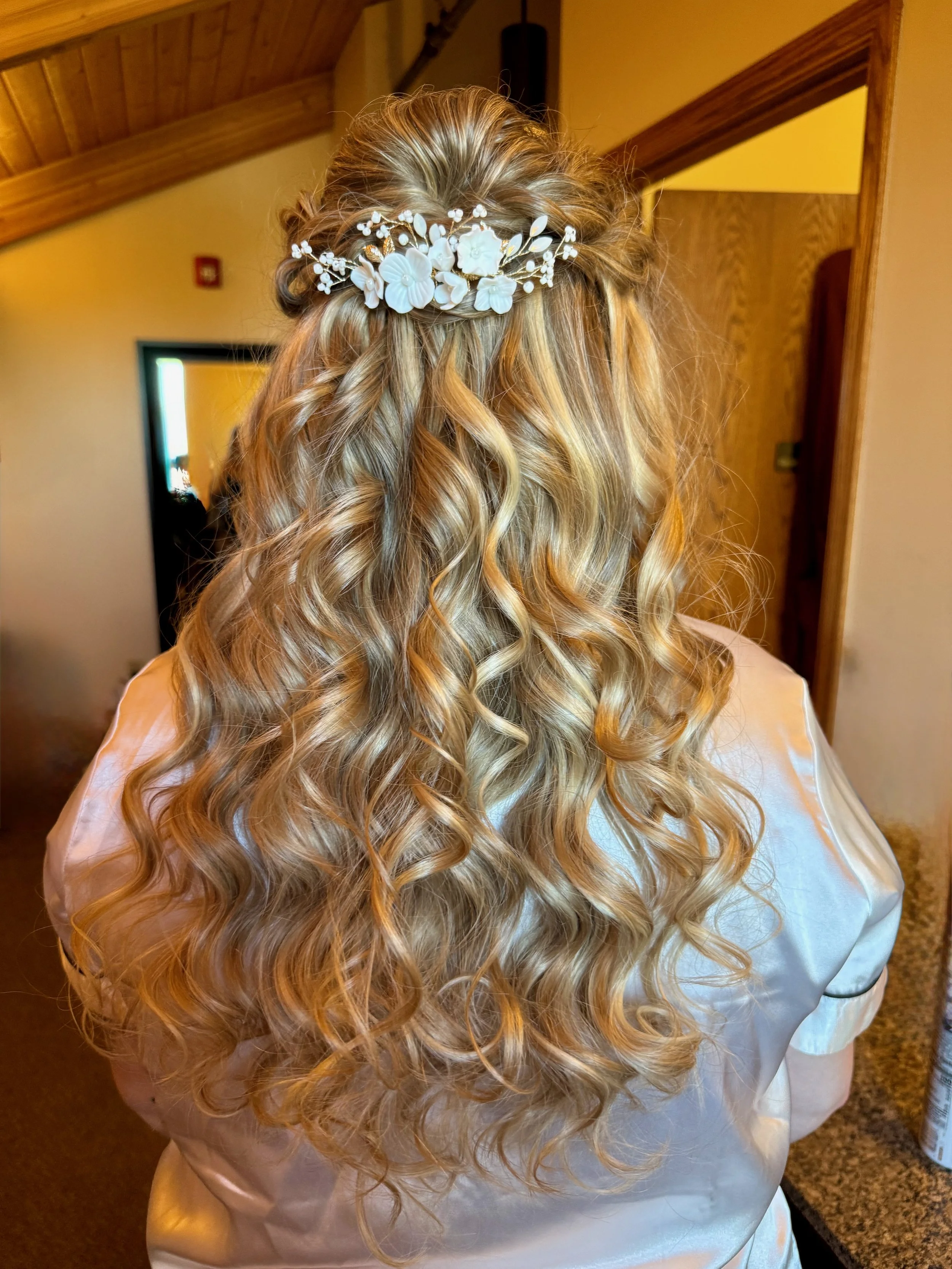 A bride with long, curly blonde hair wearing a white satin robe and a floral hair accessory with white flowers and small pearl-like accents, standing indoors near a door and wood-paneled ceiling.