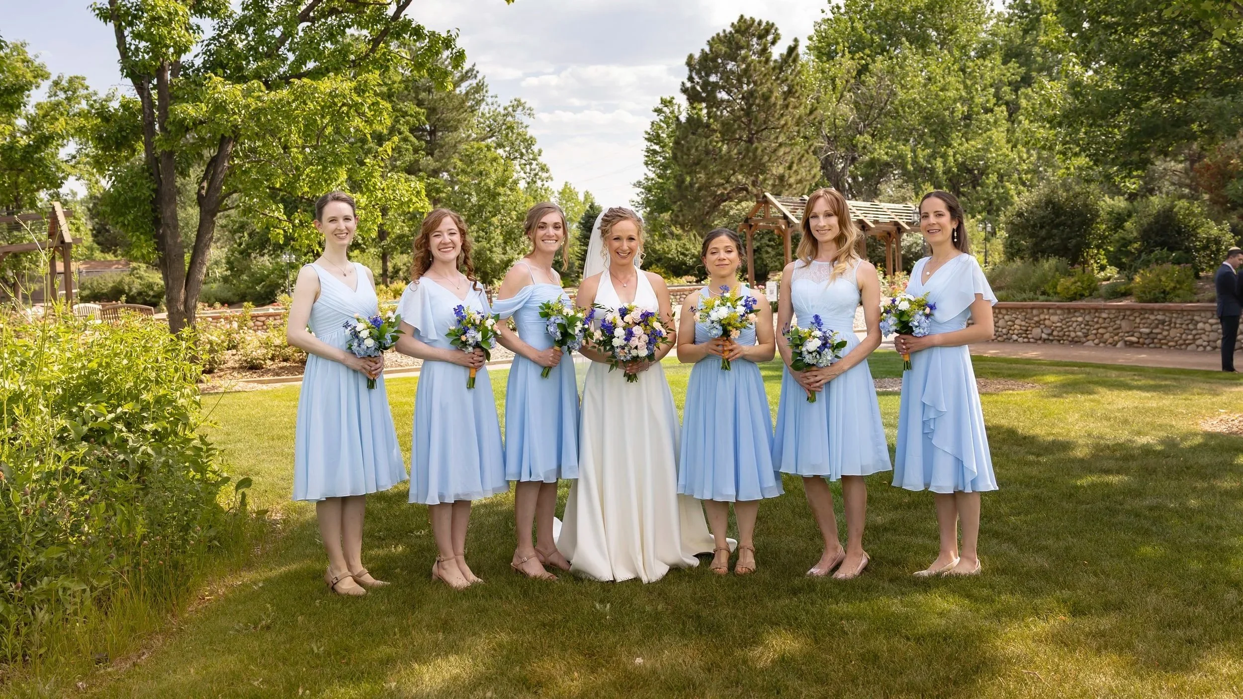 Group of seven women in light blue bridesmaid dresses standing outdoors on a grassy area, holding bouquets of flowers, with trees and a stone wall in the background, at a wedding celebration.