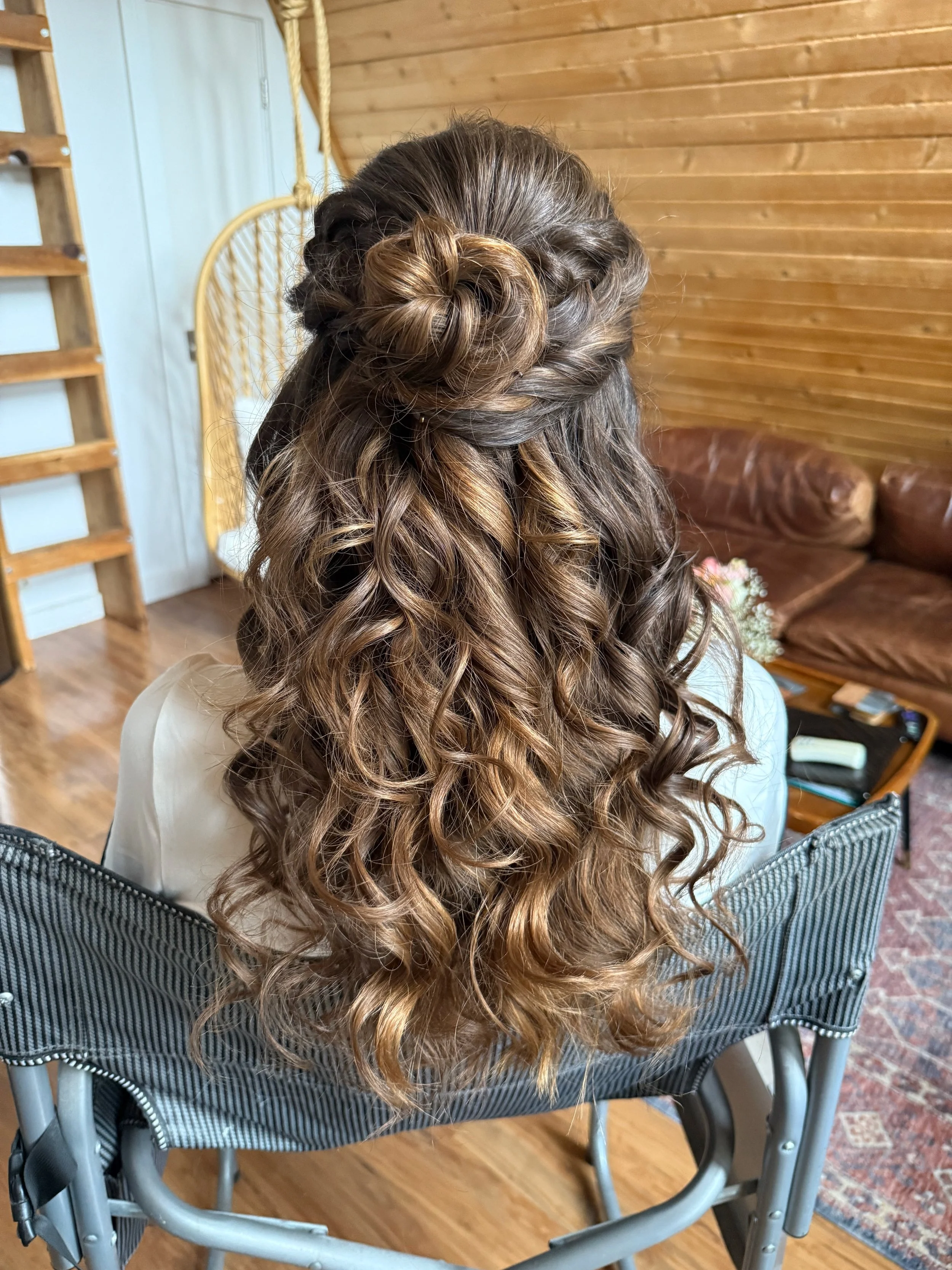 Back view of a bride with long, curly, brown hair styled in an intricate updo with a braided bun, sitting in a chair in a room with wooden walls and a brown leather couch.