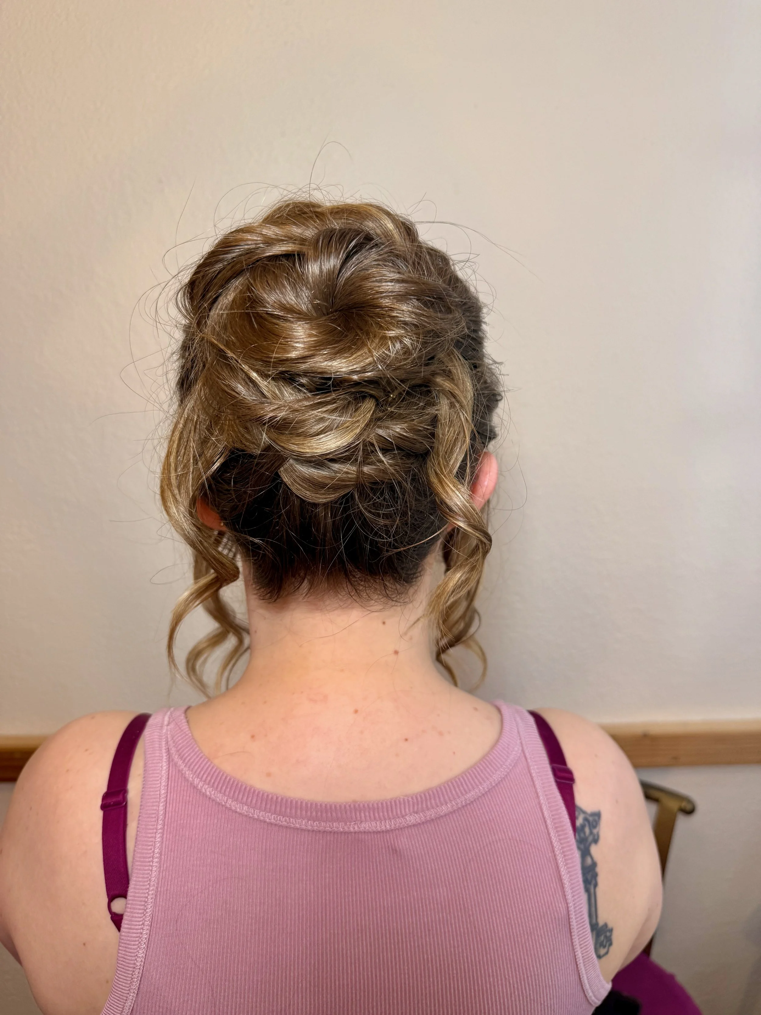 Back view of a bride with intricately styled blonde hair in an updo with curls, wearing a pink tank top with purple straps, showcasing a tattoo on her upper arm.