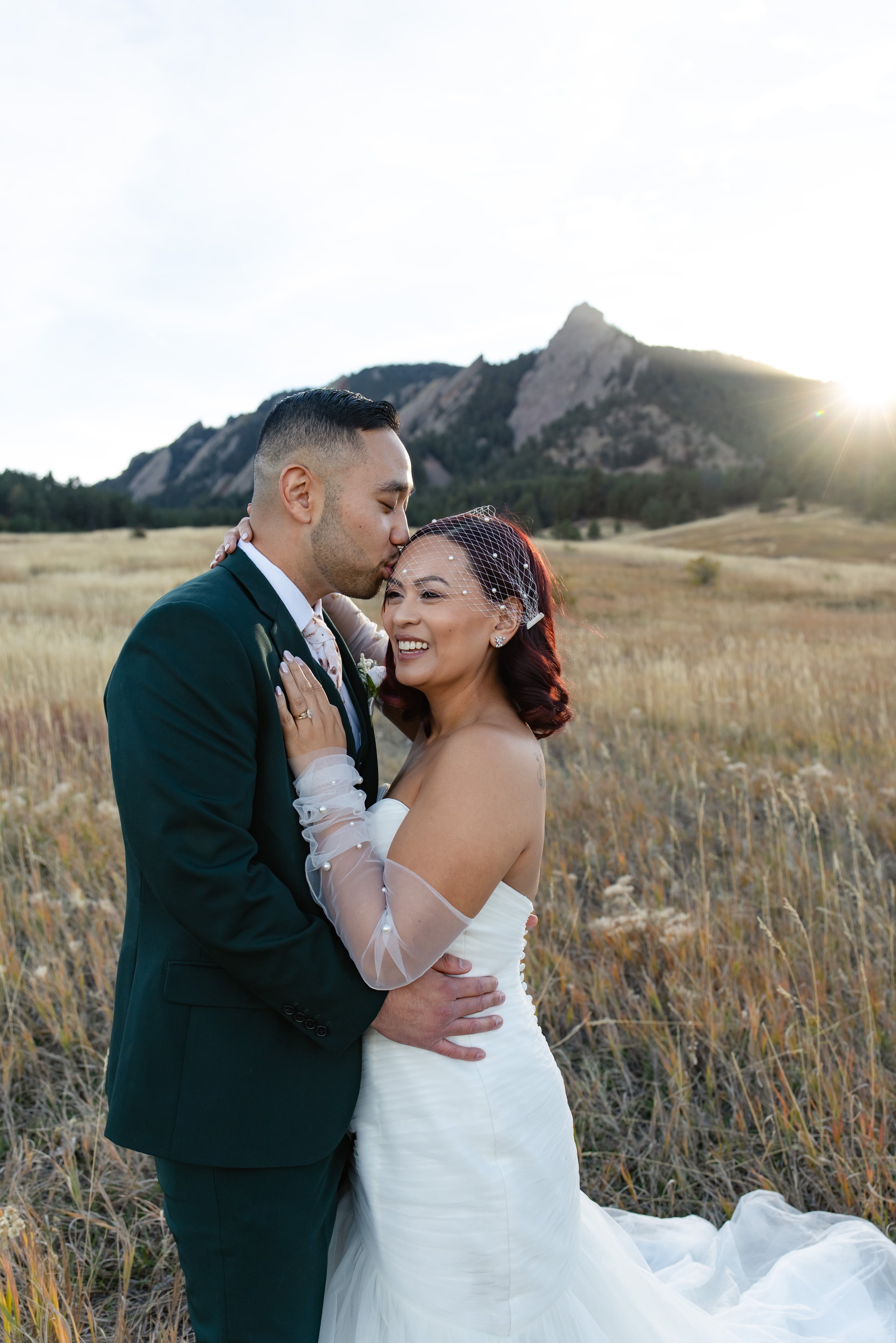 A newlywed couple in a field with mountains in the background. The groom is kissing the bride on the forehead, and they are embracing each other. The bride is wearing a white wedding dress with a veil, and the groom is wearing a dark suit. The setting is outdoors during sunset.