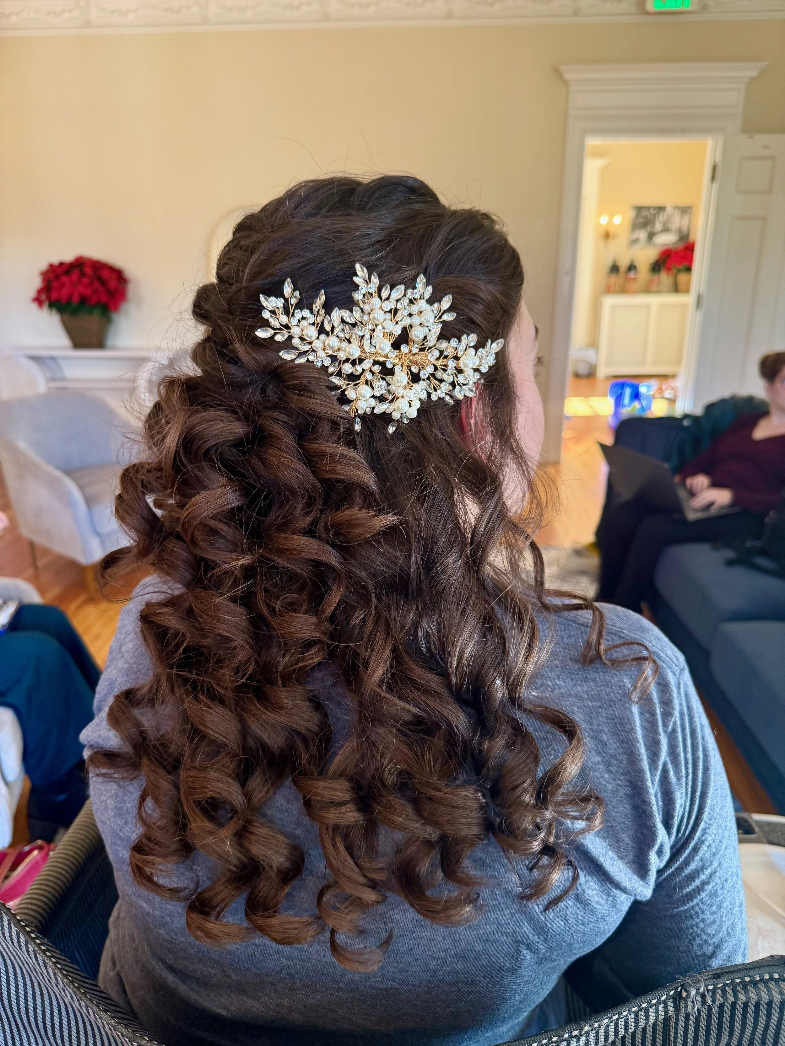 Close-up view of a bride's hair styled in loose curls, adorned with an ornate pearl and rhinestone hair accessory. The background includes a cozy room with a doorframe, a fireplace, and some holiday decorations, with a person seated on the couch.