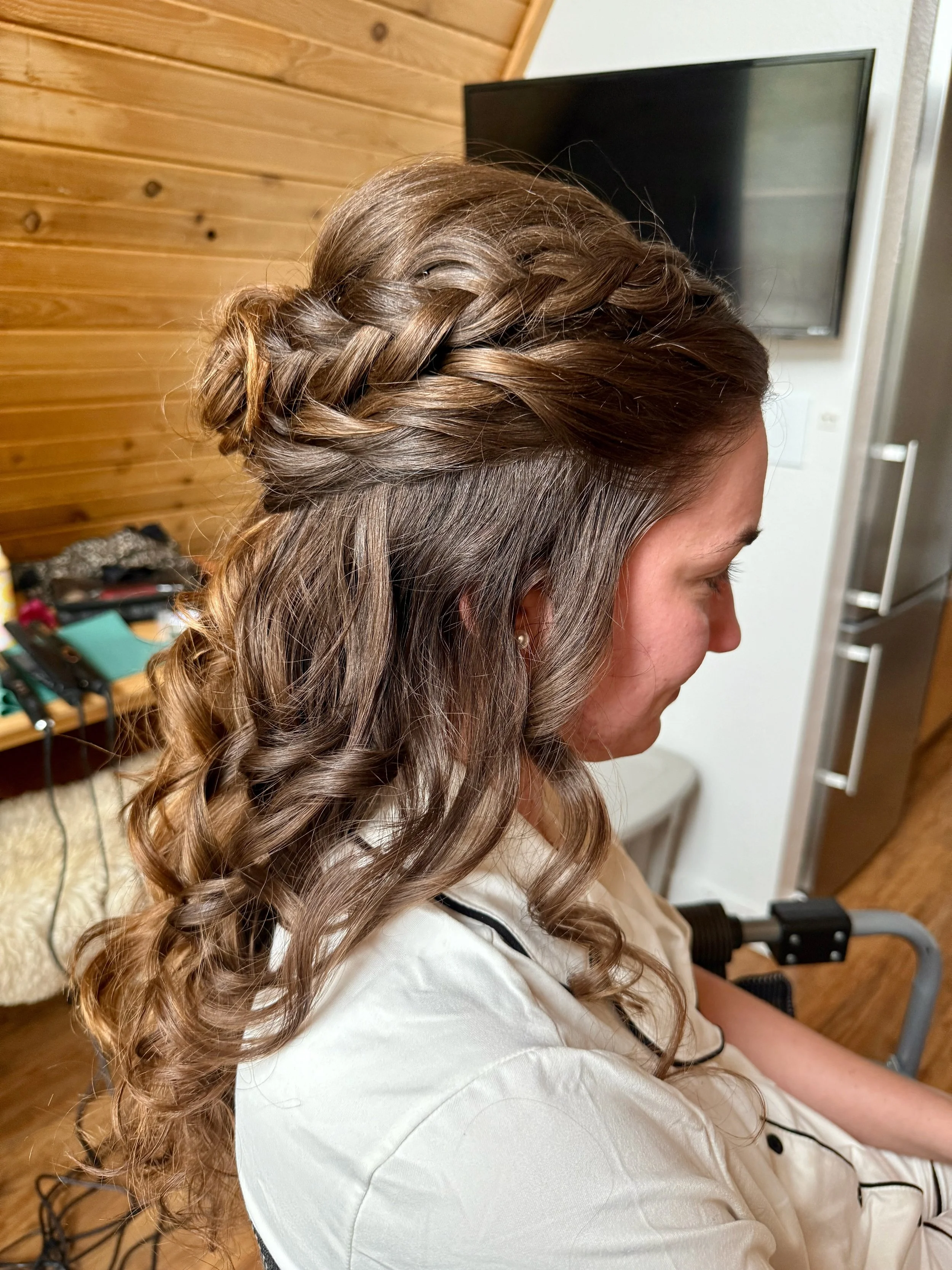 A bride with long, curly hair styled in a braided crown, sitting indoors near a television and wooden wall.