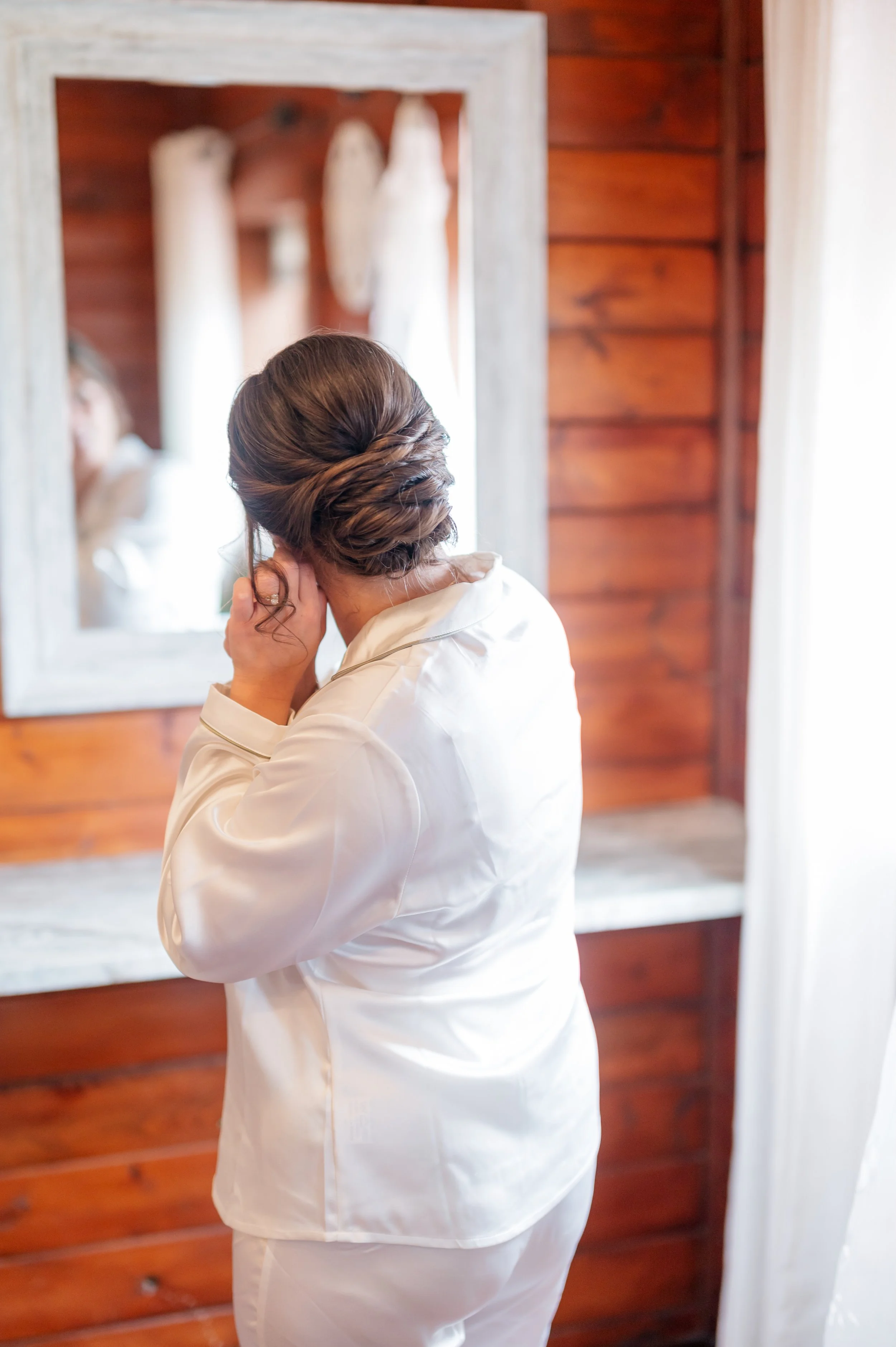 A bride with brown hair styled in an elegant updo, wearing a white satin robe, appears to be looking at herself in a mirror.