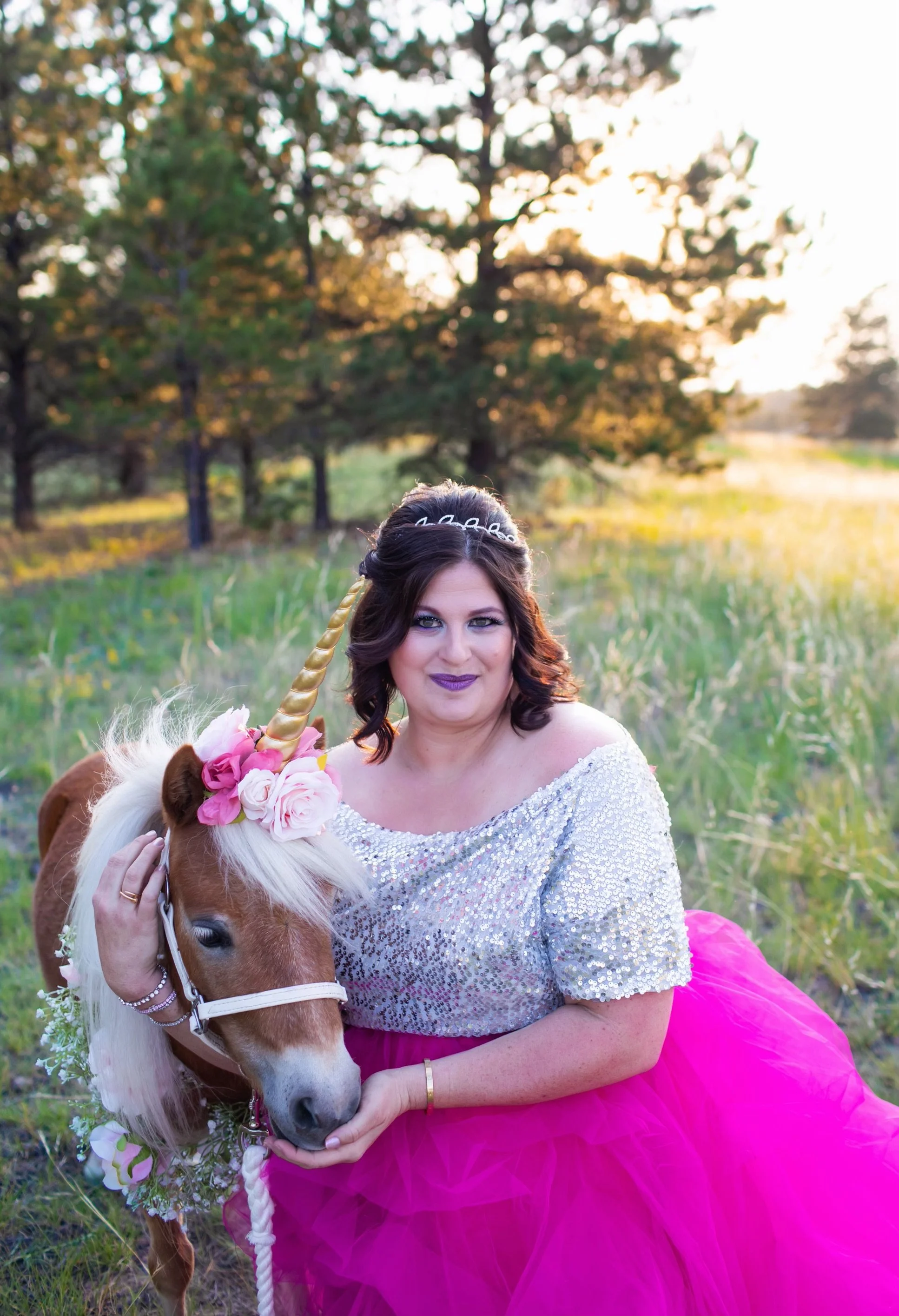 A woman dressed in a sparkly silver top and bright pink tutu, with dark hair styled with a tiara, holding a small brown and white pony with a floral crown and a golden horn, in a green outdoor field during sunset.