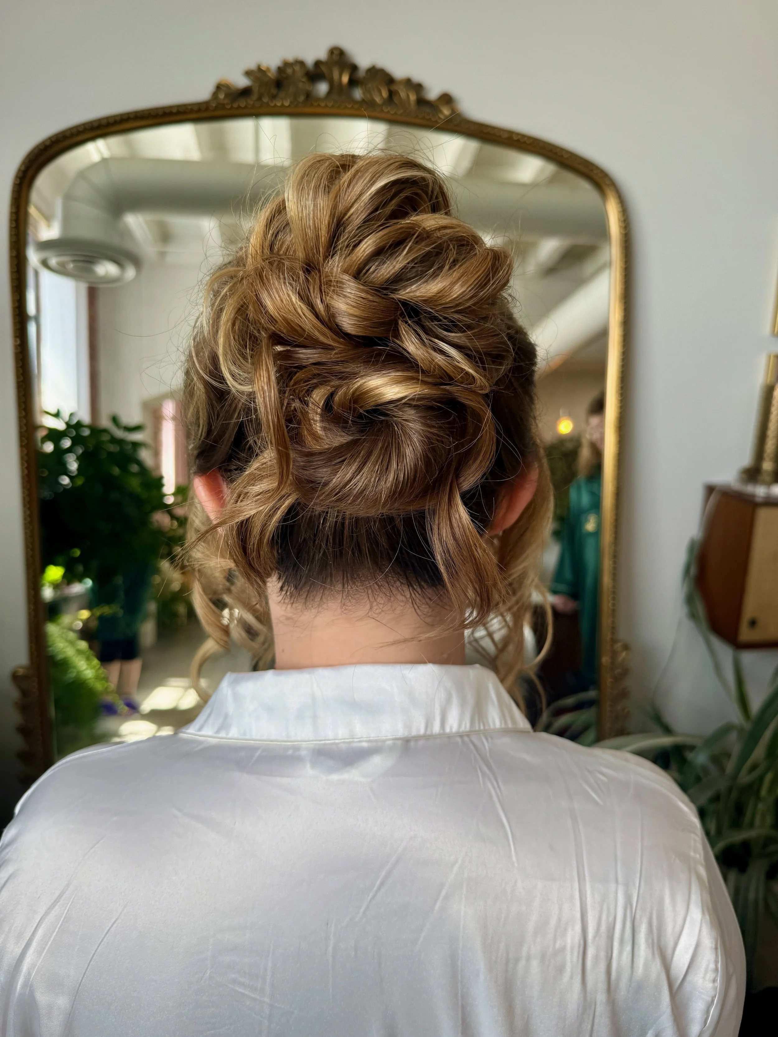 The back of a bride's head with styled, curled, light brown hair, seen in a mirror with a decorative gold frame, reflecting an indoor space with plants and another person in the background.