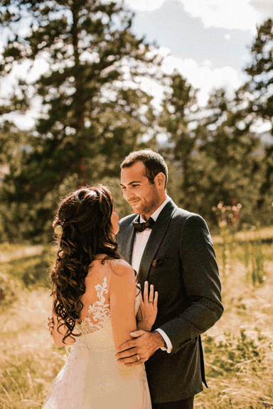 A bride and groom standing close outdoors in a grassy field with trees in the background during daytime, looking at each other lovingly.
