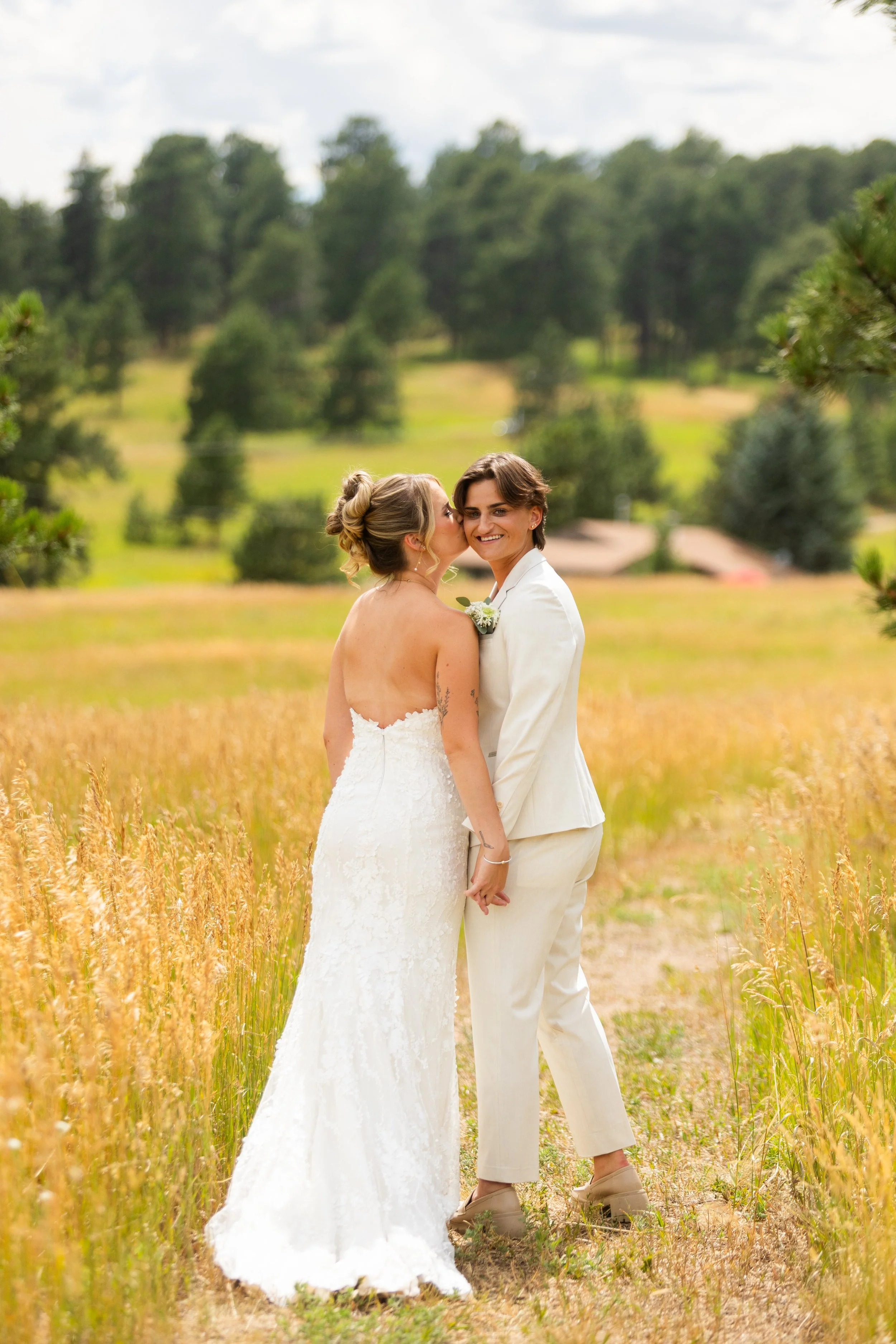 A same-sex couple in wedding attire standing in a field of tall, golden grass with trees and a cloudy sky in the background. One woman is kissing the other on the cheek, who is smiling at the camera.