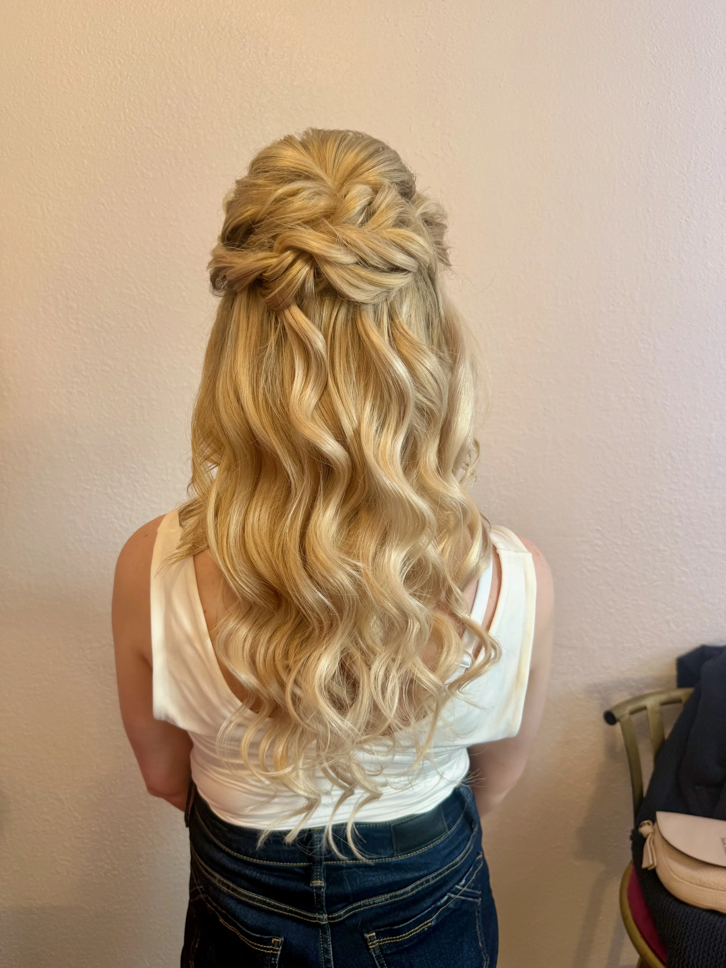 Back view of a bride with long, wavy, blonde hair styled with a twisted braid crown, wearing a cream-colored sleeveless top and dark jeans, standing against a beige wall.