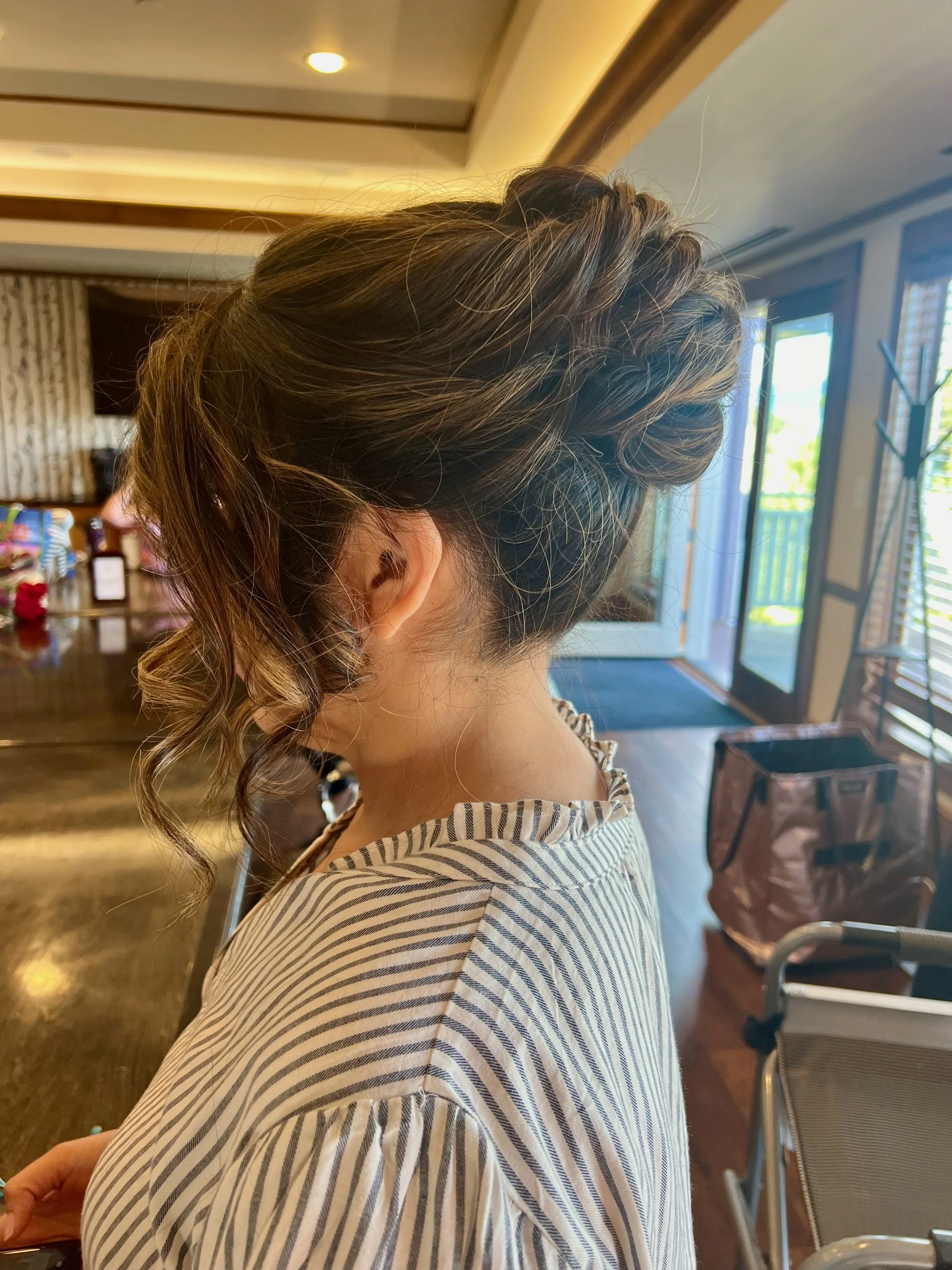 A bride with hair up with loose curls, wearing a striped shirt, sitting indoors near a window with sliding glass doors, in a warmly lit room.