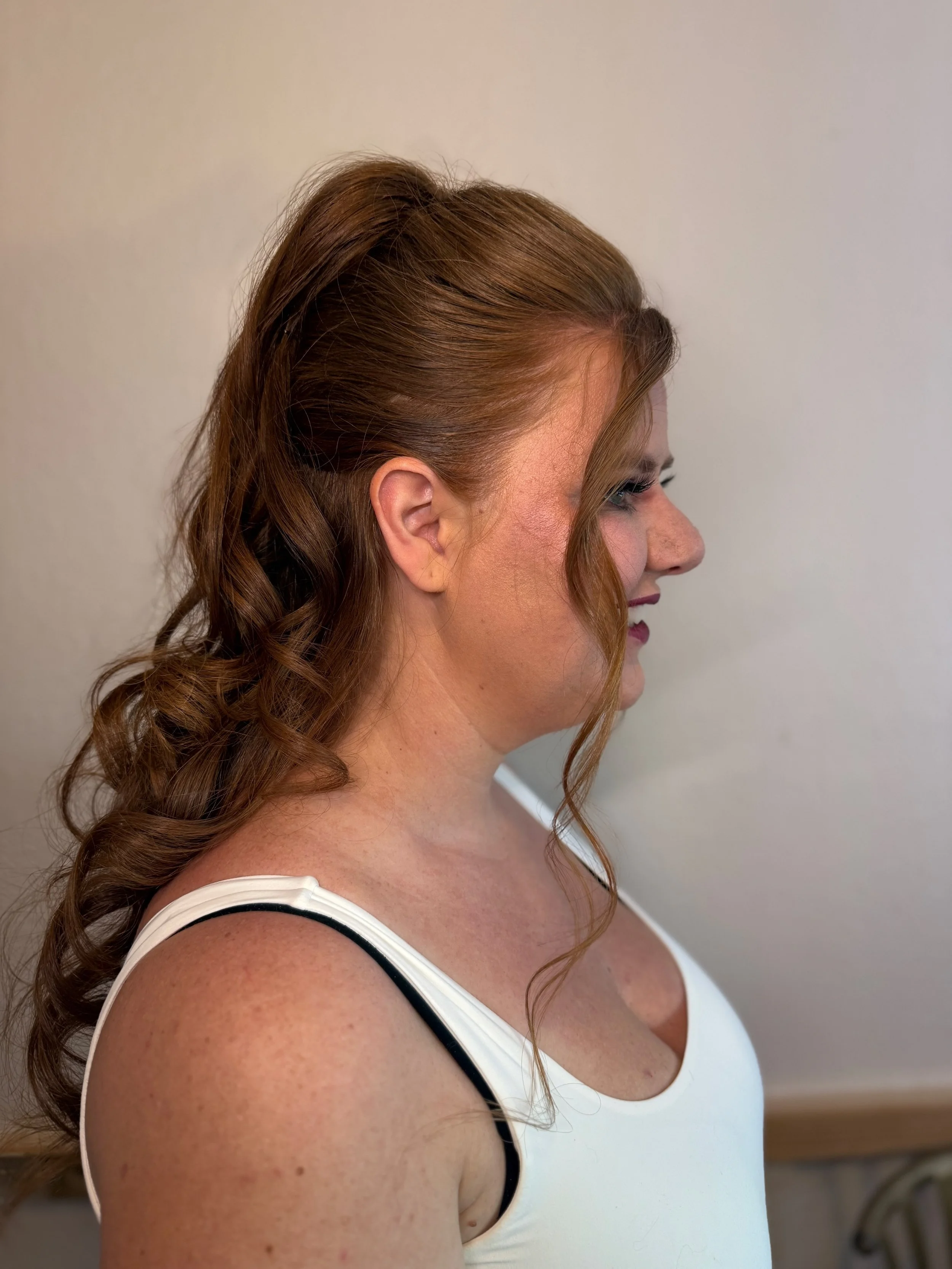 Profile of a bride with long, wavy, auburn hair styled in loose curls, wearing a white tank top with black straps, standing against a plain wall.