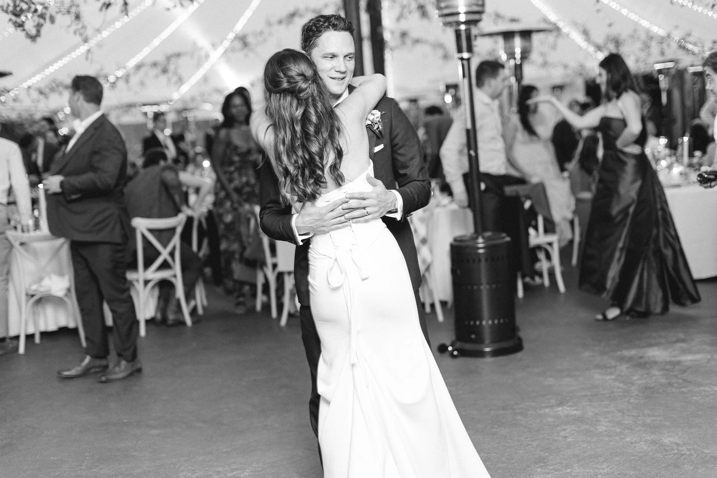 A couple dancing closely at an elegant event, surrounded by seated guests and decorated with string lights. The bride has a half-up hair style. 