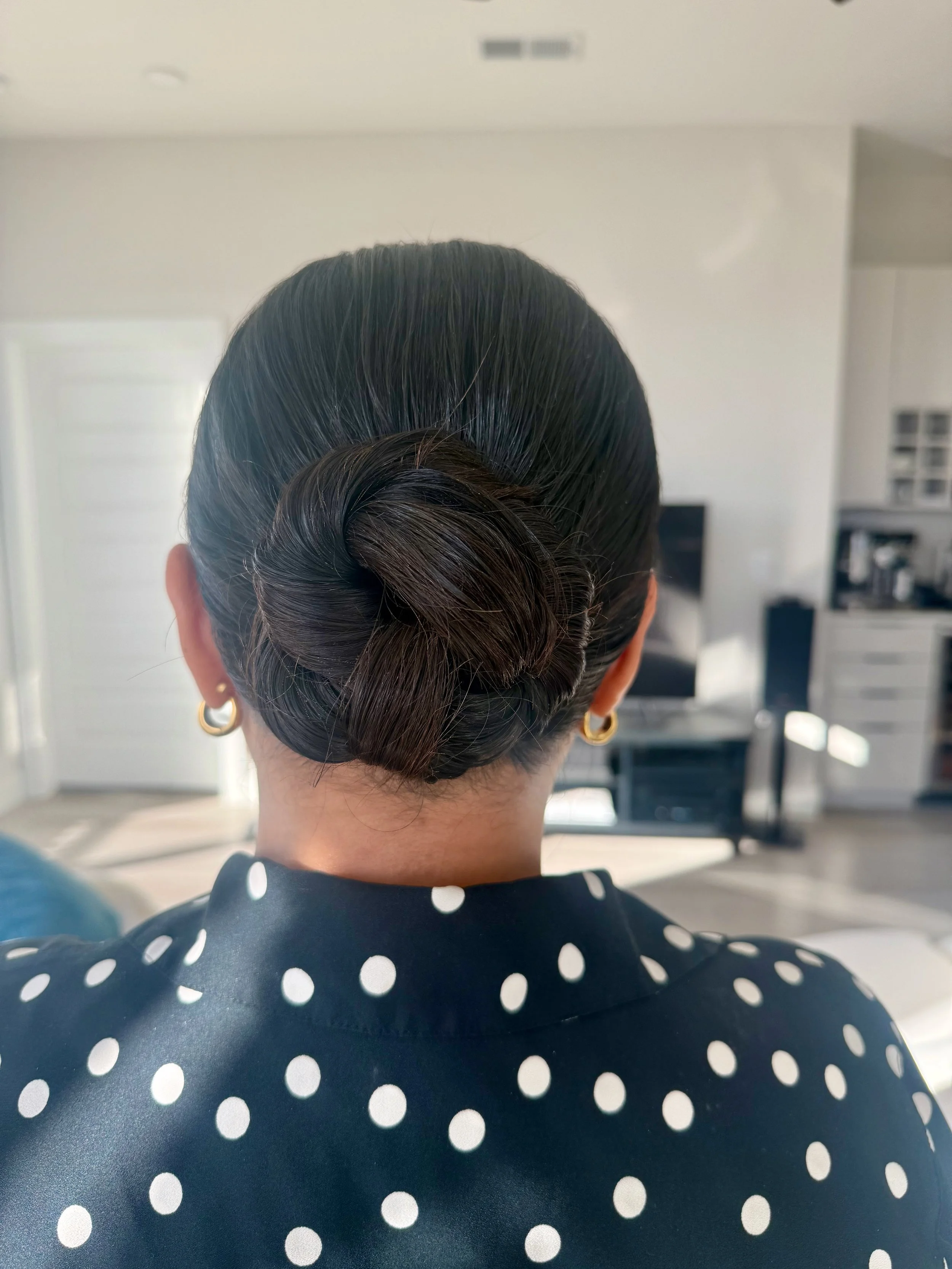 Back view of bride with dark hair styled in a vintage twist, wearing a navy blue dress with white polka dots and gold hoop earrings, sitting in a bright living room.