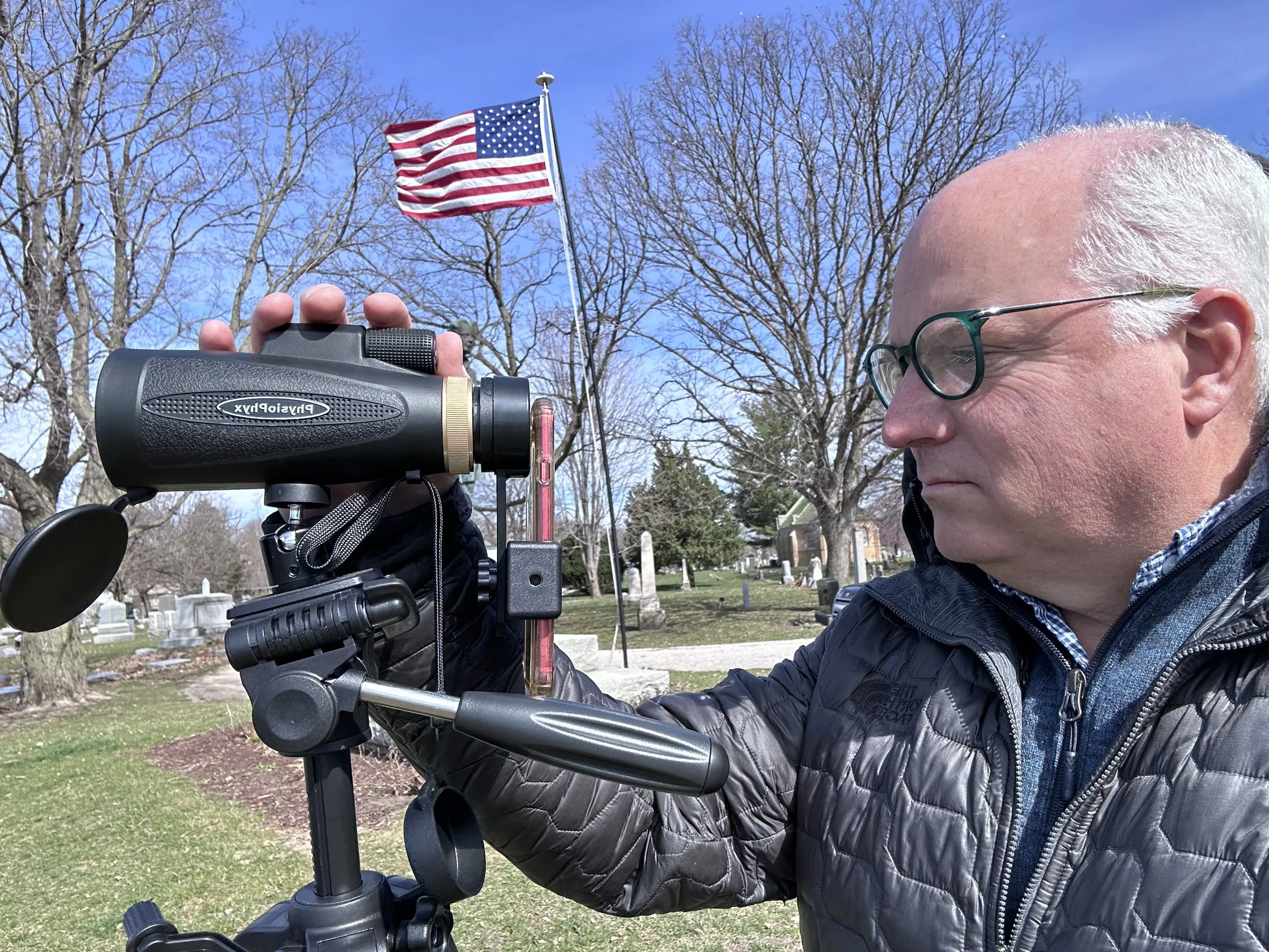 A man with glasses and white hair holding a device mounted on a tripod outdoors in a park or cemetery with leafless trees and an American flag on a flagpole in the background.
