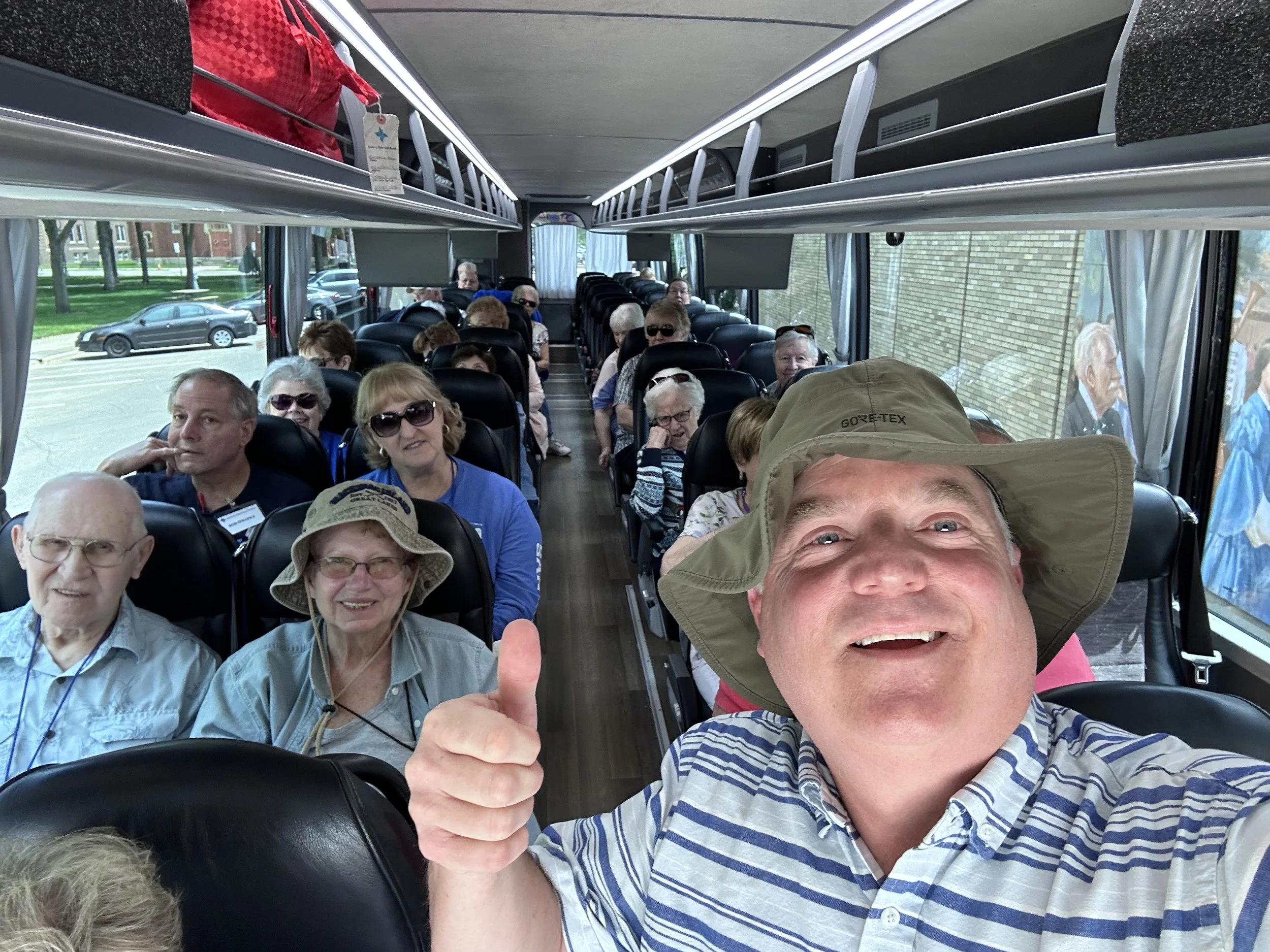 A group of senior citizens on a bus, with the man in the front taking a selfie and giving a thumbs-up, many wearing sunglasses and hats, seated in black leather seats, with large windows showing the street outside.