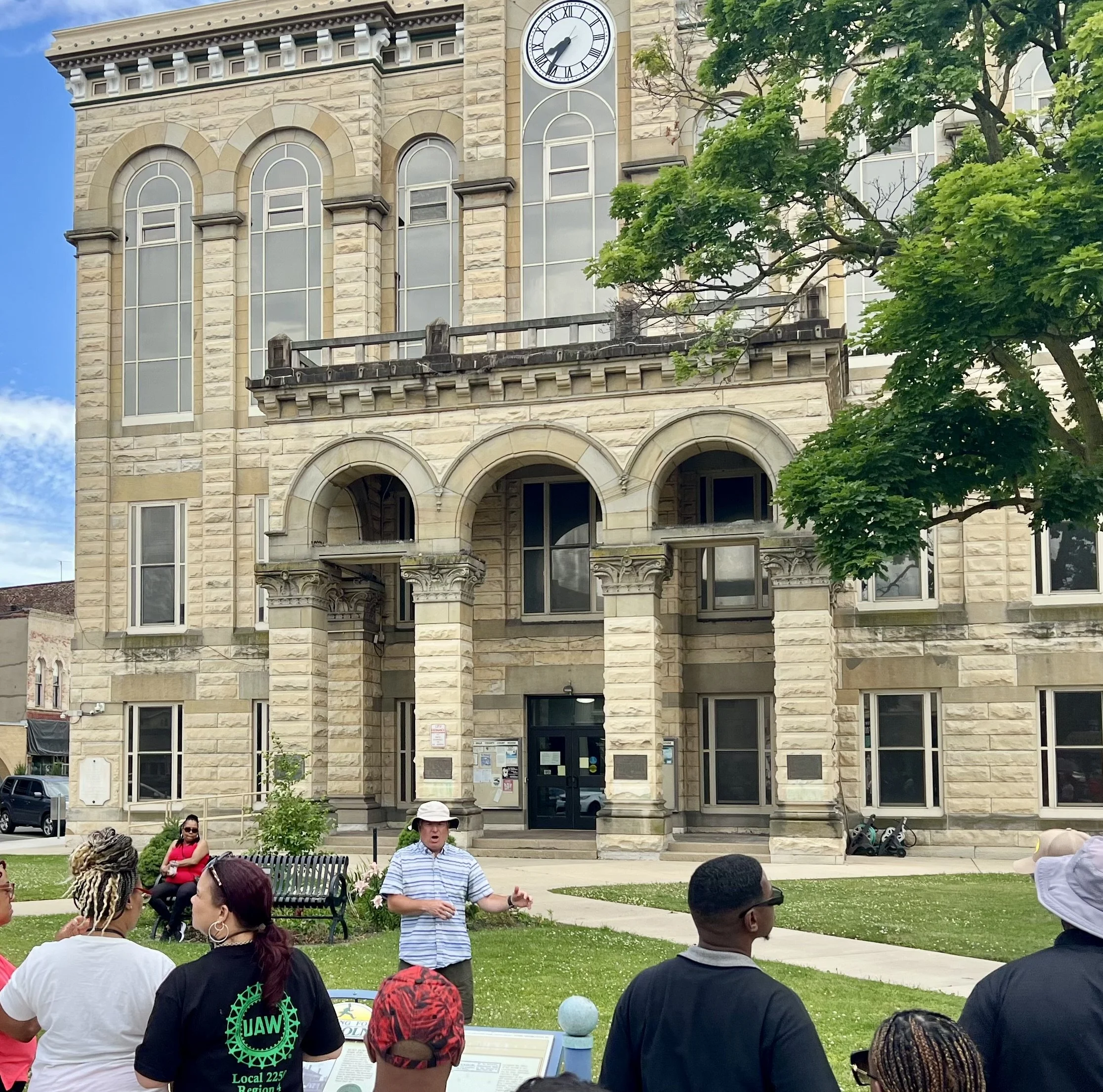 A group of people attending a tour or outdoor gathering in front of a historic stone building with a clock tower. The building features arched windows and classical architectural details, with a small grassy area and trees in the scene.