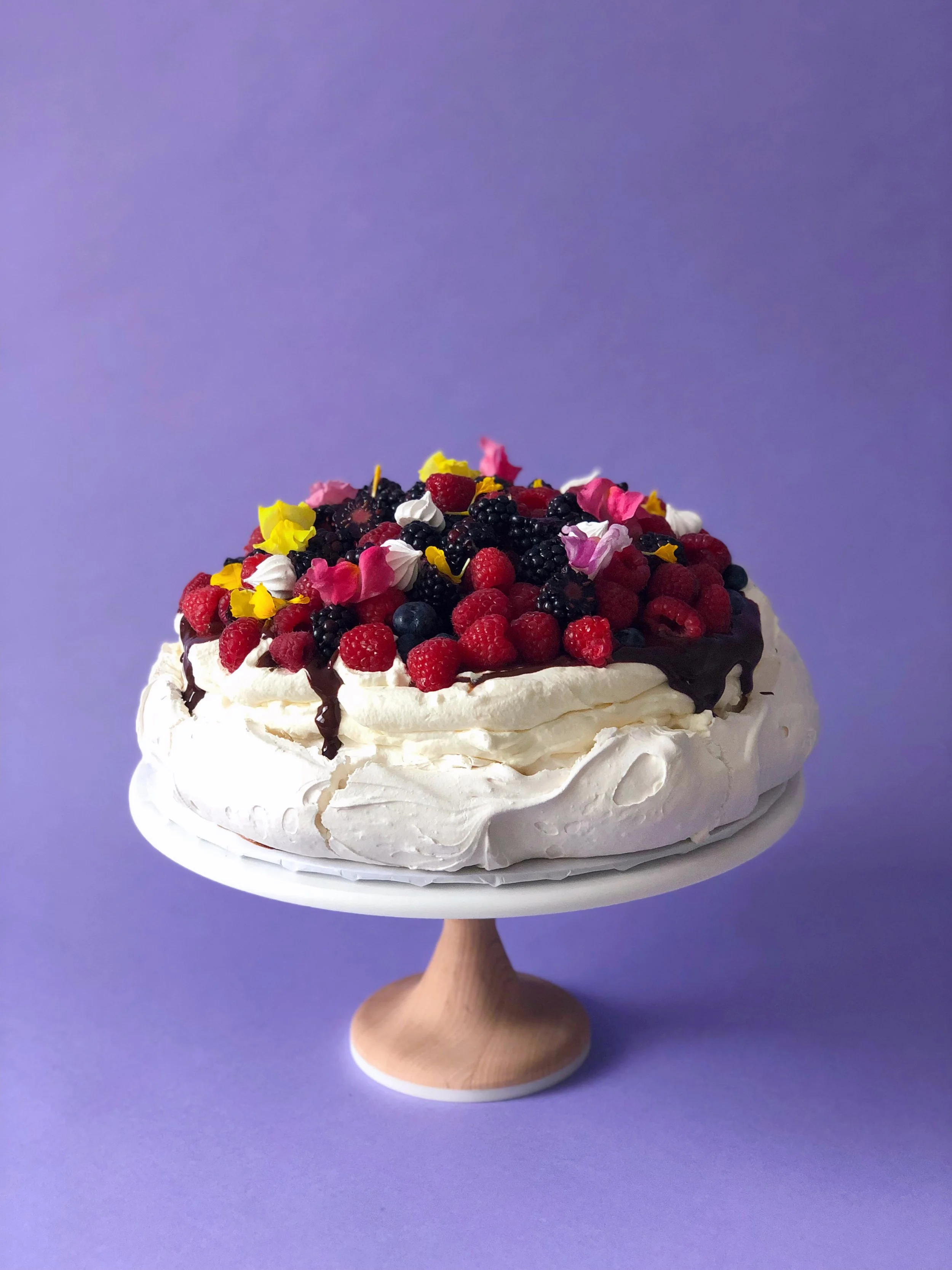 Crisp meringue shell filled with vanilla whipped cream, drizzled with dark chocolate ganache, and topped with seasonal berries, mini meringues, and edible florals.