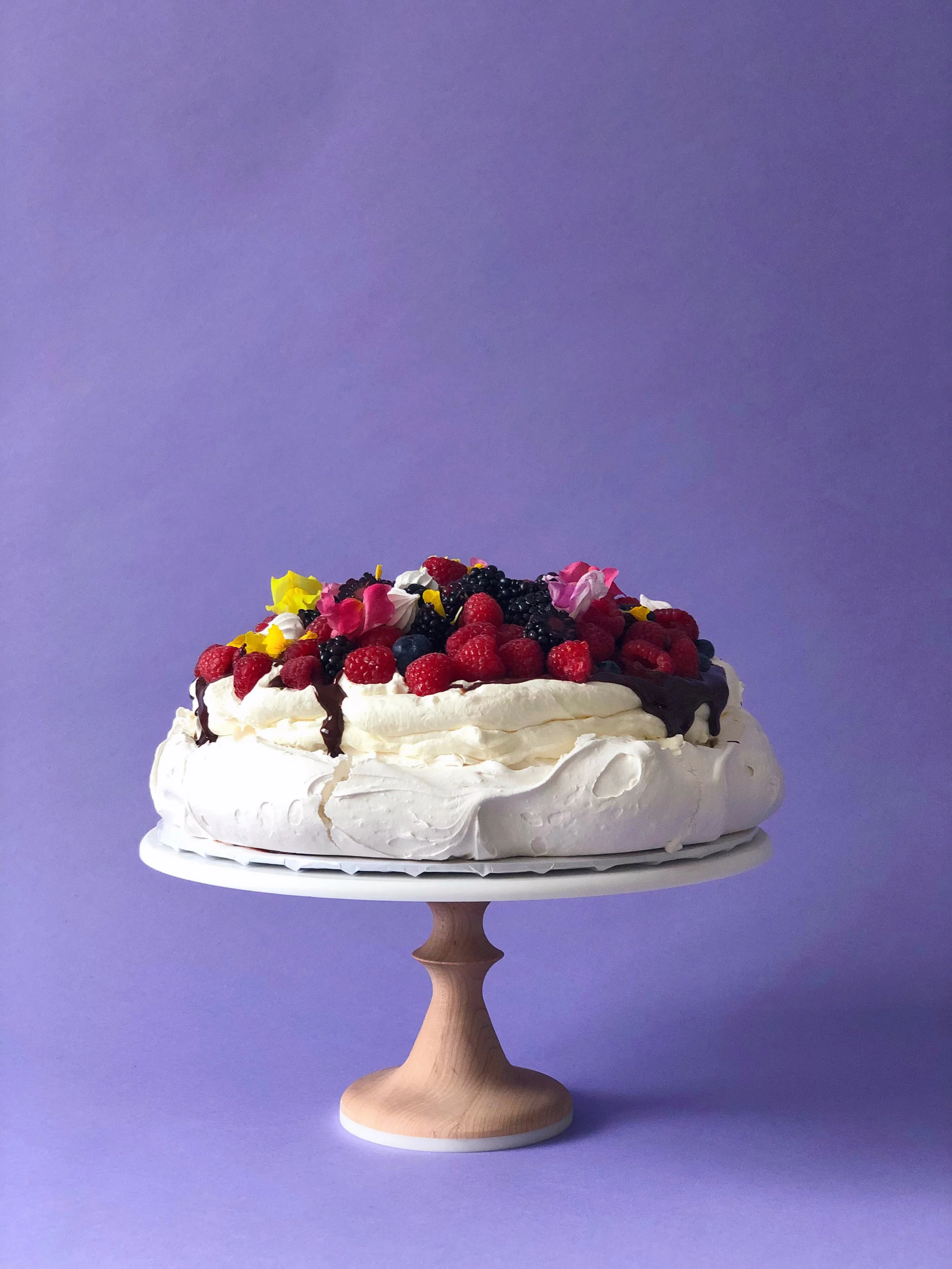 Crisp meringue shell filled with vanilla whipped cream, drizzled with dark chocolate ganache, and topped with seasonal berries, mini meringues, and edible florals.