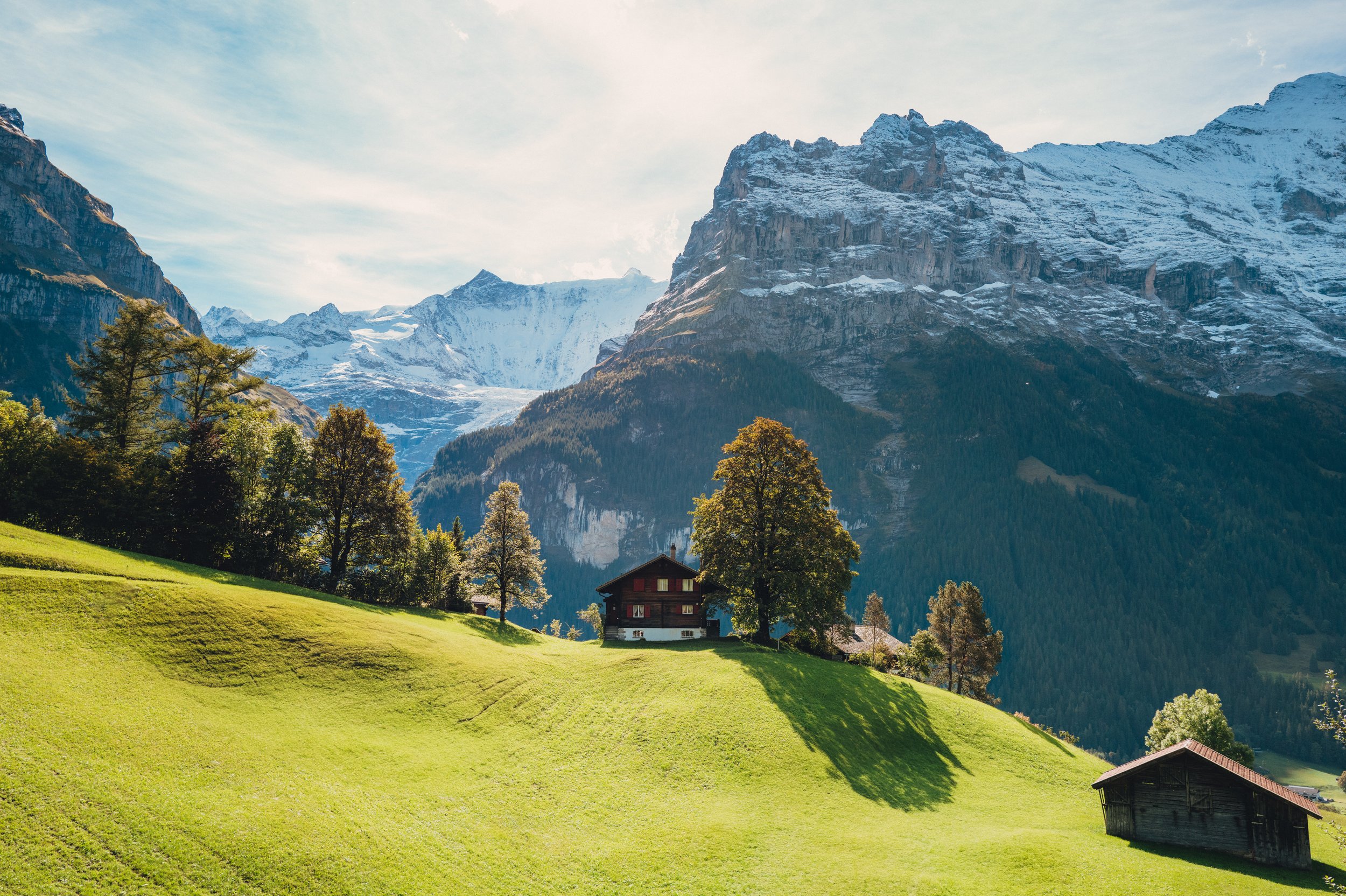 Green rolling hills with trees and houses at the foot of snow-capped mountains under a partly cloudy sky.