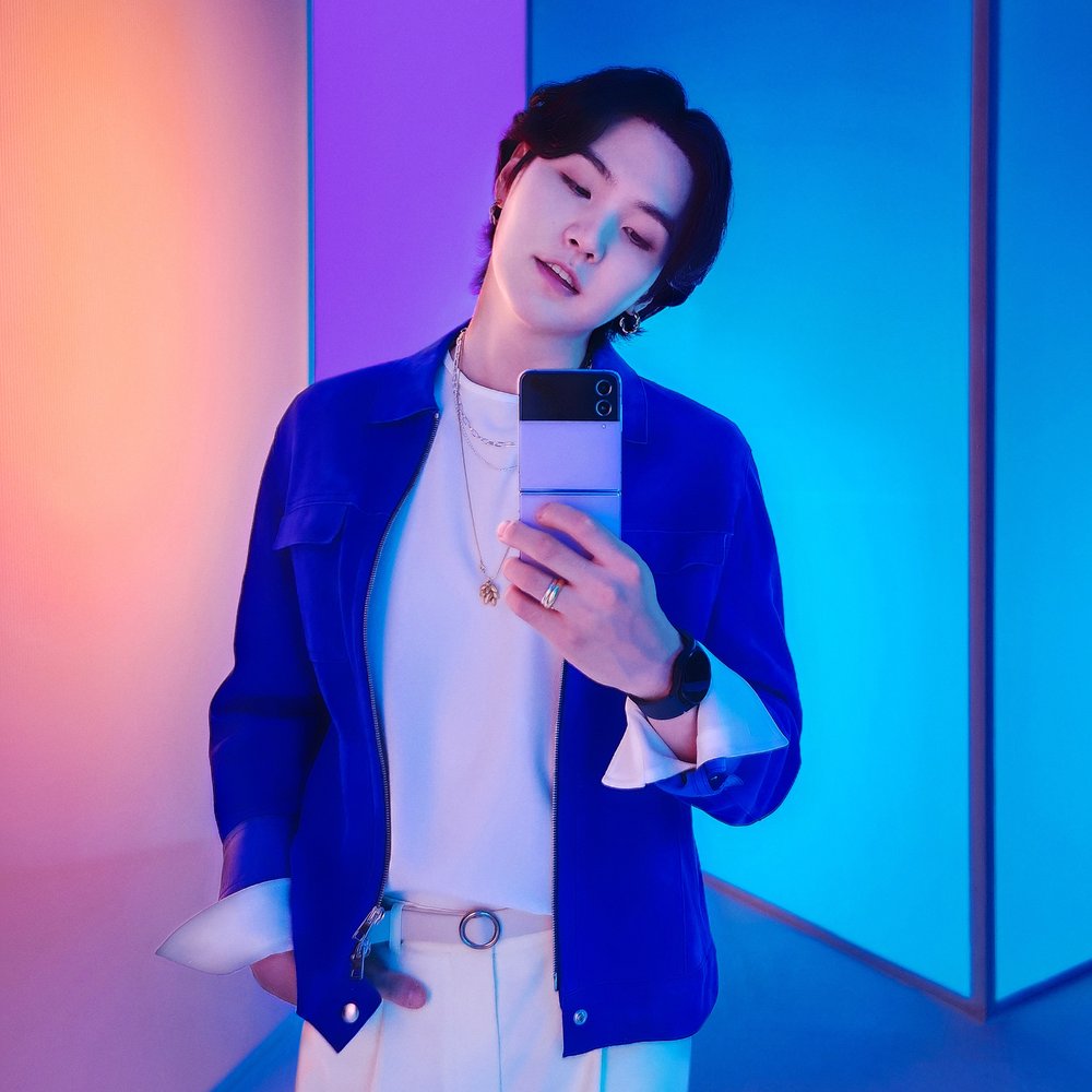 Suga in blue &amp; purple holding the Galaxy ZFlip4. 