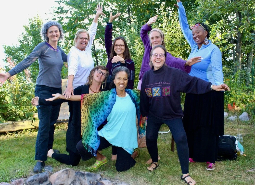 Group of nine women smiling and posing outdoors in a garden, with some raising their arms and one kneeling in front, surrounded by greenery.