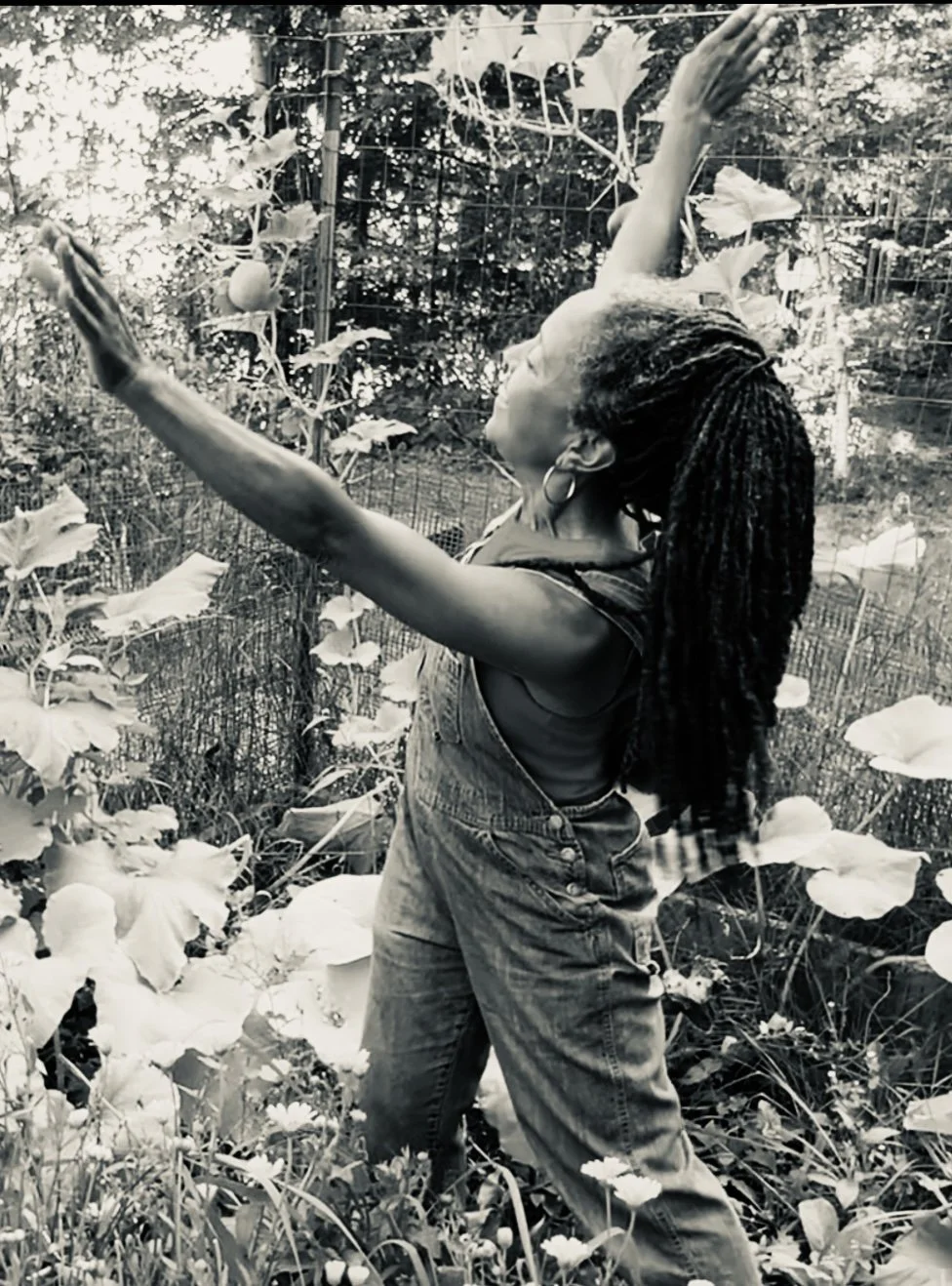 A woman with dreadlocks and hoop earrings stands with arms outstretched among large-leafed plants in a garden or natural setting, smiling with eyes closed.