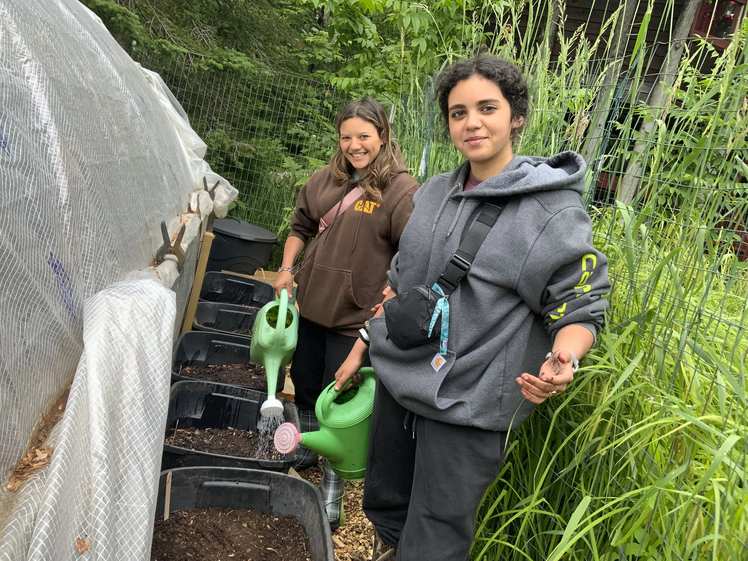 Two young women gardening outdoors, watering plants in black containers lined up on a raised surface near a greenhouse with mesh covering. One woman is smiling, holding a green watering can, wearing a brown hoodie. The other woman is holding a small plant, wearing a gray hoodie, standing among green plants.