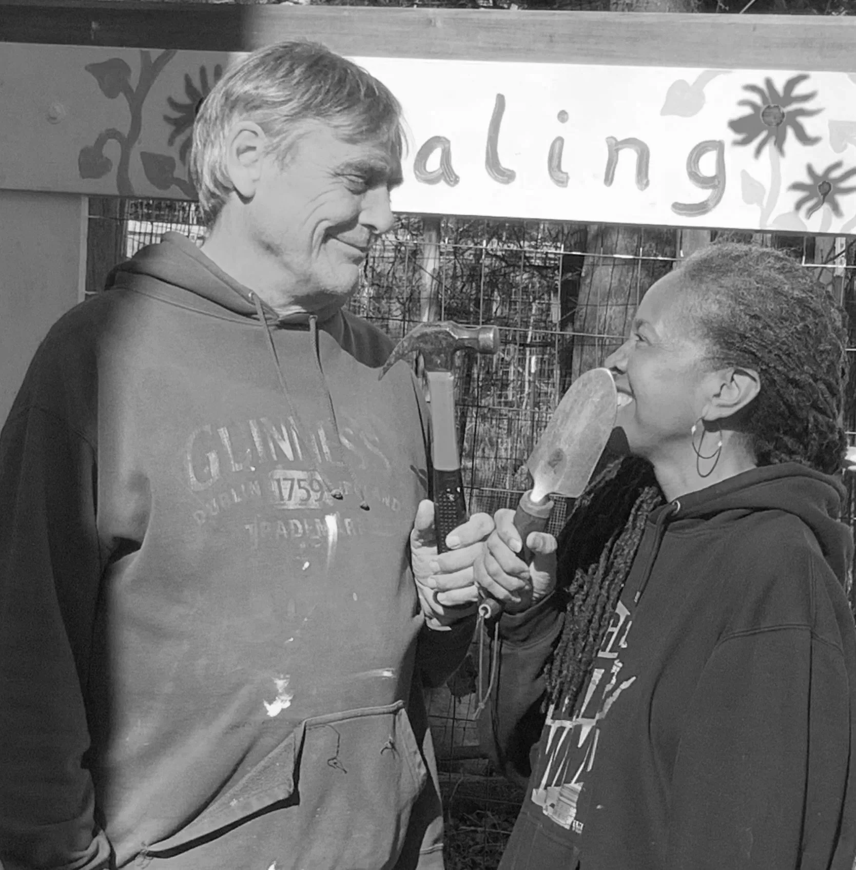 A man and woman smiling at each other, holding gardening tools, in front of a sign that reads 'Valing'.