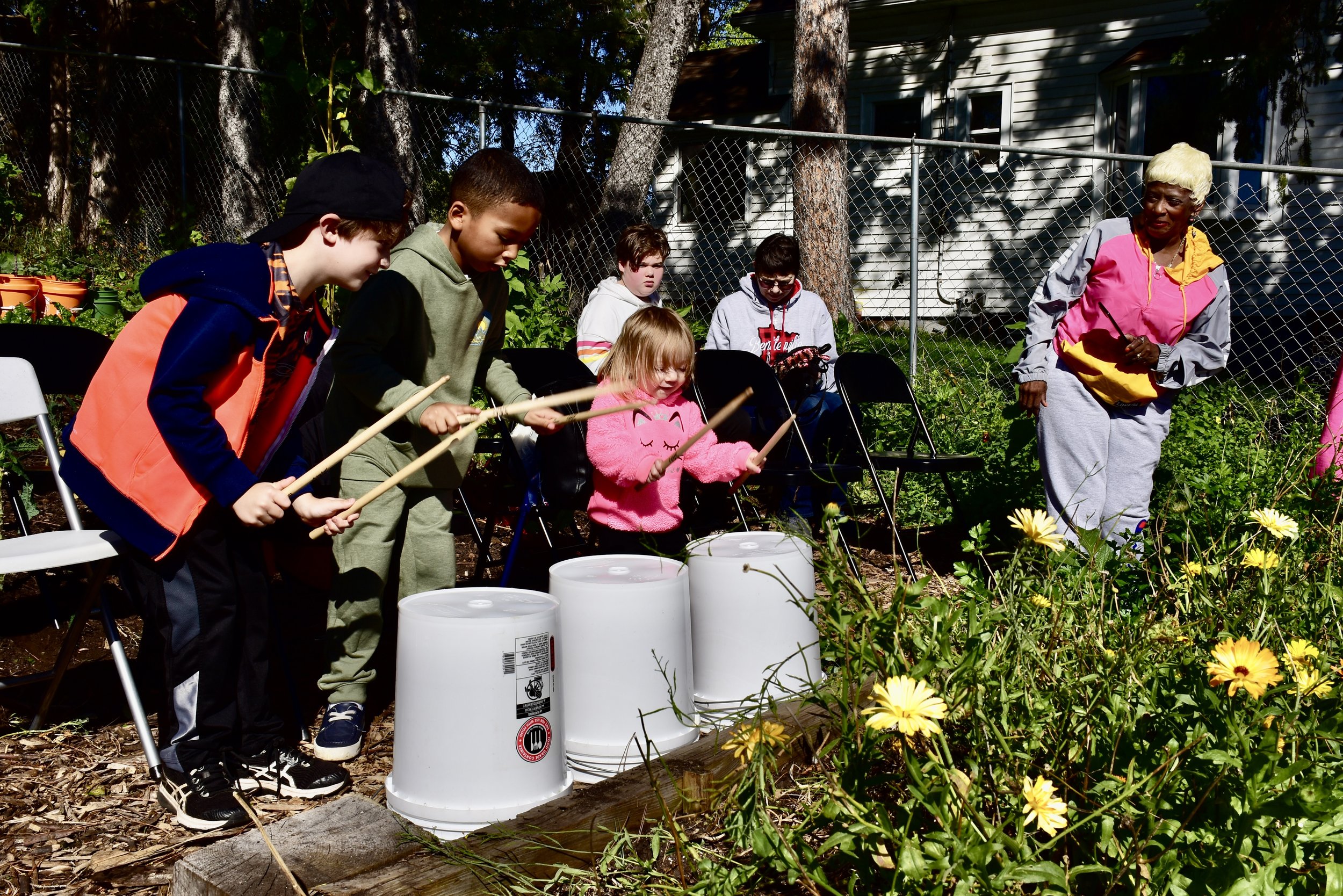 Children and an adult in a garden, with some children playing musical instruments, others seated, surrounded by flowers and greenery.