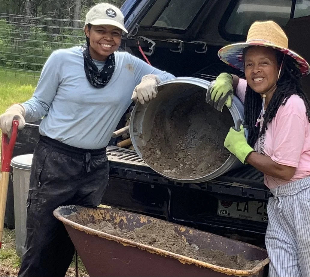 Two women are smiling and working together outdoors, pouring dirt or concrete from a large round metal container into a wheelbarrow. They are wearing work gloves and hats, and are standing next to a vehicle with its hatch open.