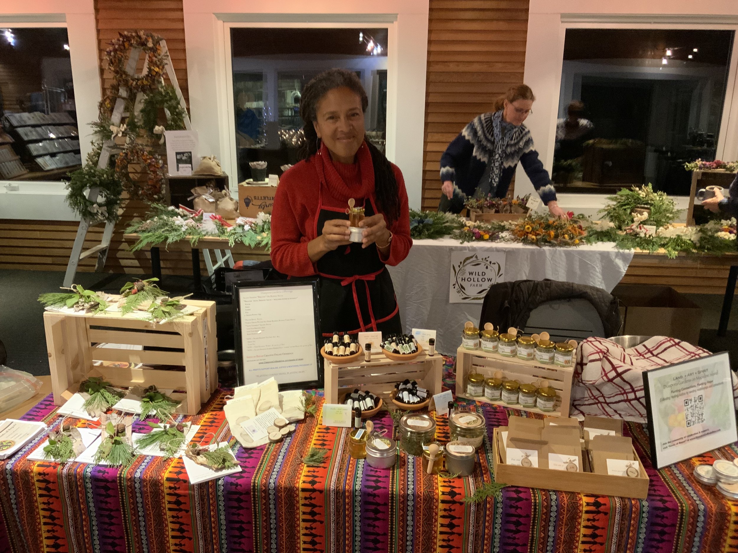 A woman in a red sweater and black apron holding a small item at a craft stall decorated for the holiday season, with another woman arranging crafts behind her. The table is covered with a colorful patterned cloth displaying various handmade items, jars, and signs.