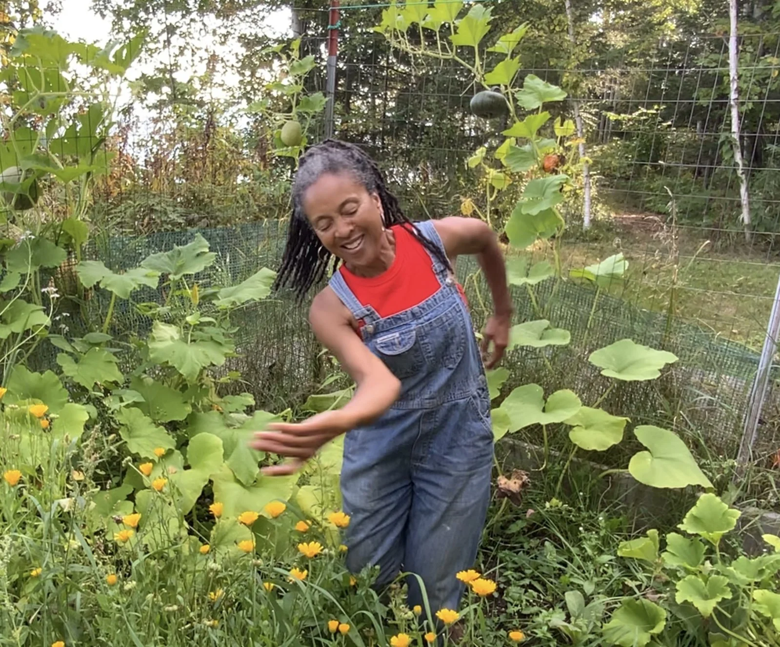 A woman with dreadlocks, wearing a red shirt and denim overalls, smiling and dancing in a lush garden with green foliage and yellow flowers.