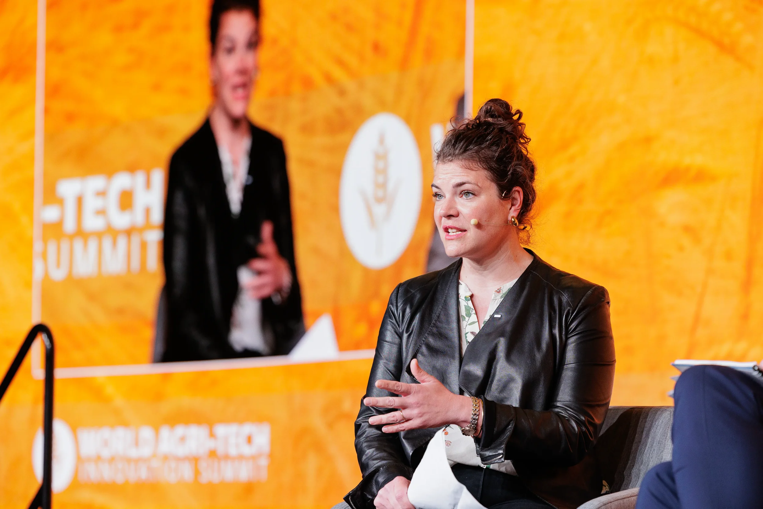 Female panelist in leather jacket with earpiece speaks on stage at Chicago corporate industry conference