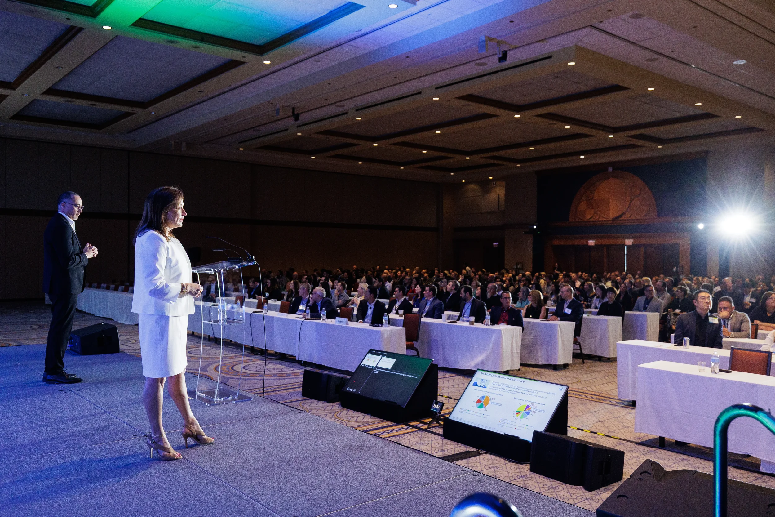 Presenter at clear podium addresses full ballroom audience seated at long tables at Chicago corporate meeting