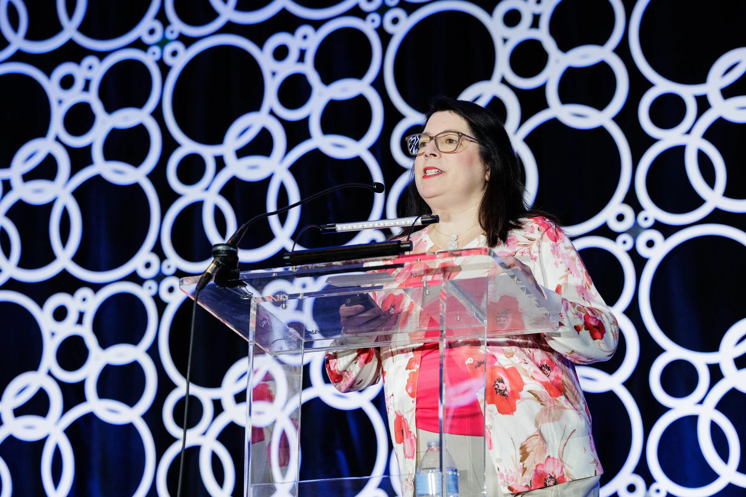 Speaker in floral cardigan presents from clear podium with circle backdrop at Chicago corporate event