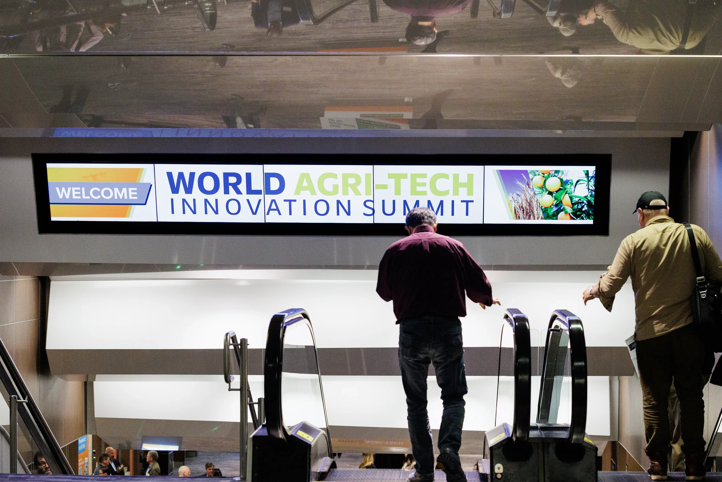 Two attendees ride escalator past large branded welcome screen at Chicago corporate industry conference