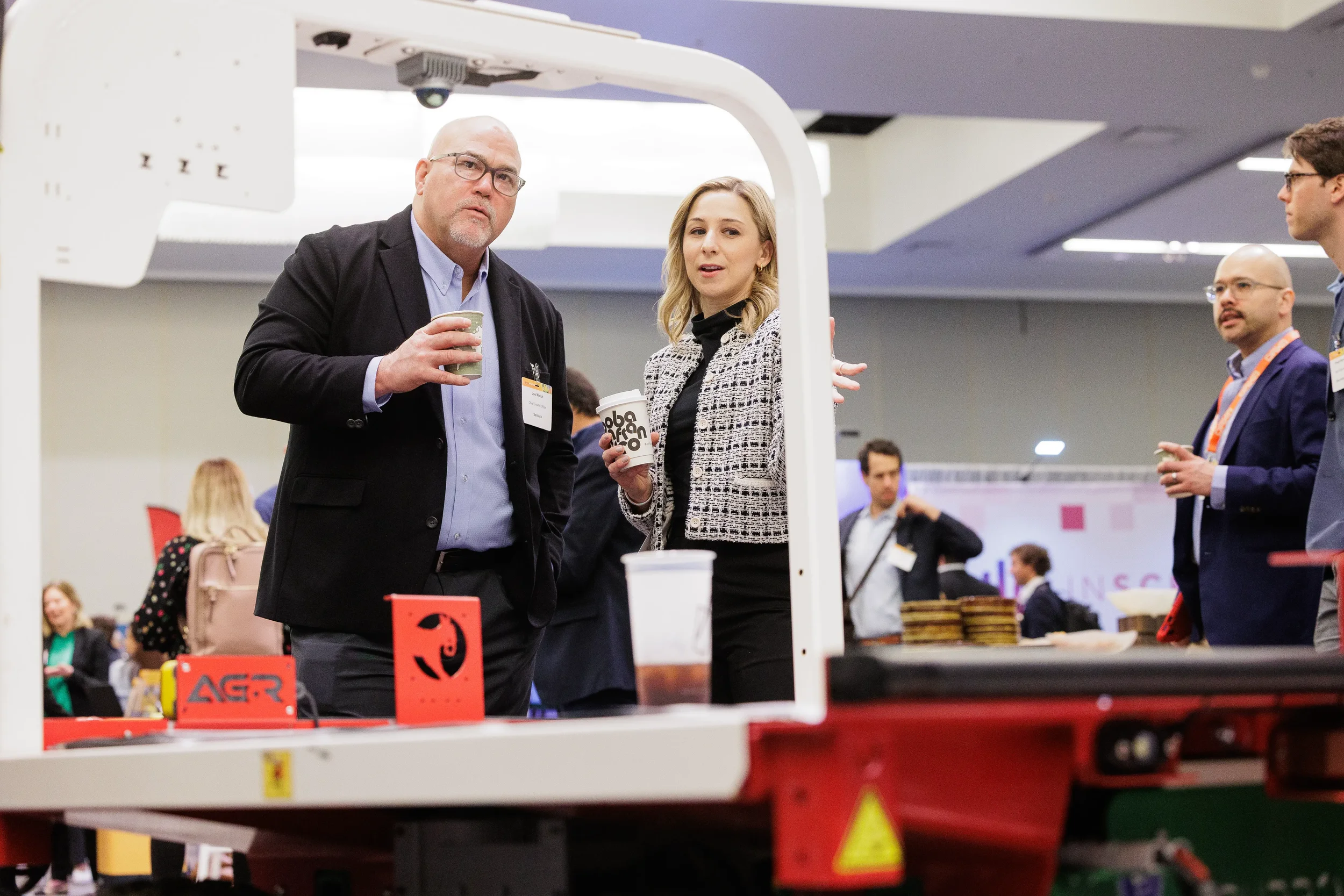 Two badged attendees with coffee examine agricultural robot on Chicago industry conference trade show floor