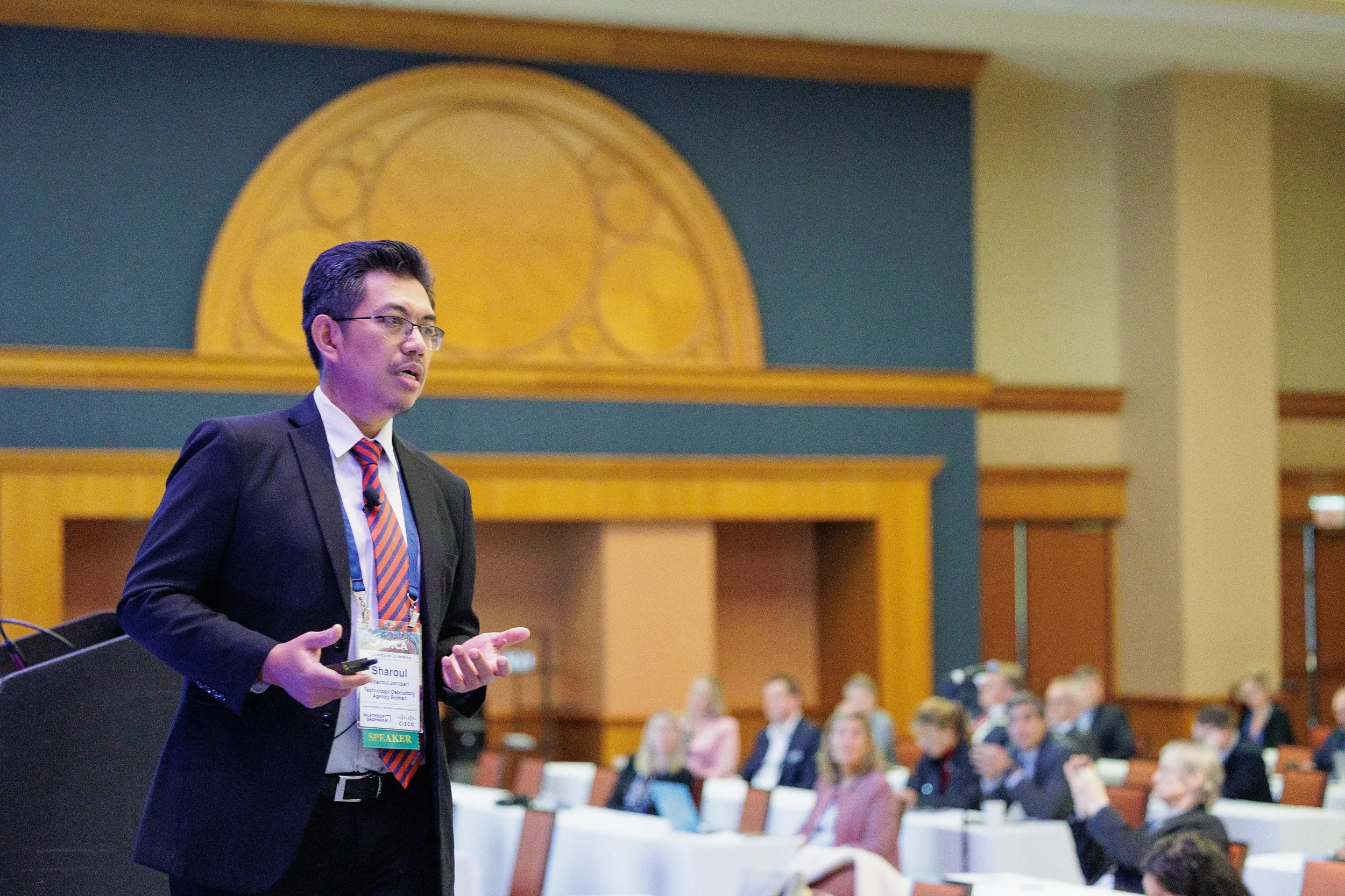 A speaker in a suit and glasses presents to an audience in a Chicago conference room with blue and gold decor.