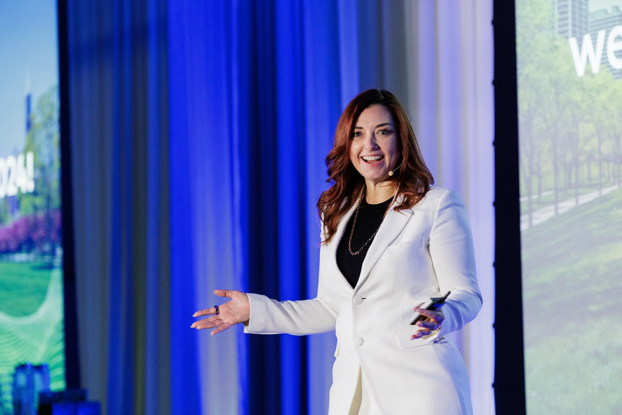 Smiling speaker in white suit gestures with open hand on stage at Orlando corporate conference