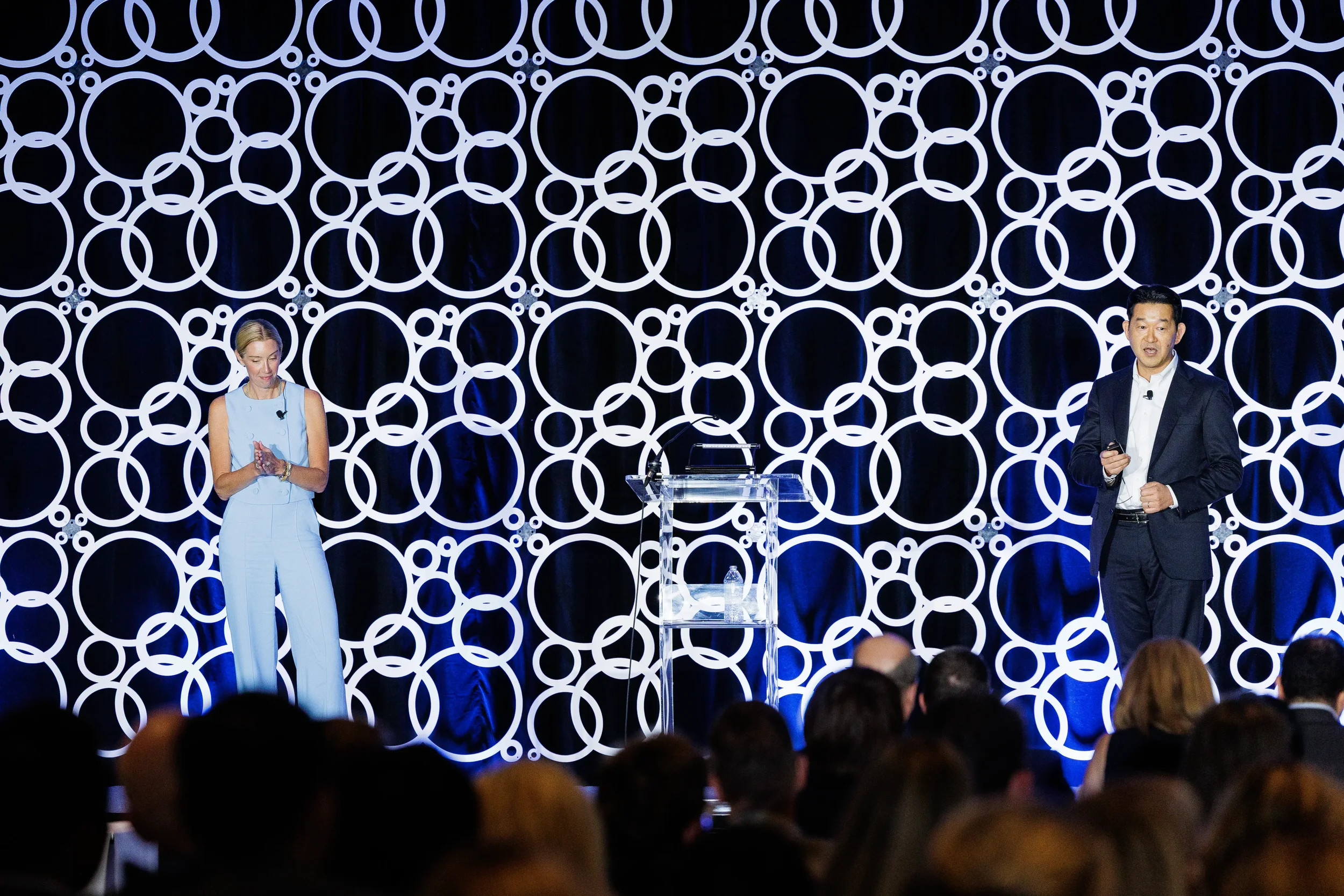 Two presenters on stage beside clear podium with circle-patterned backdrop at Chicago corporate meeting